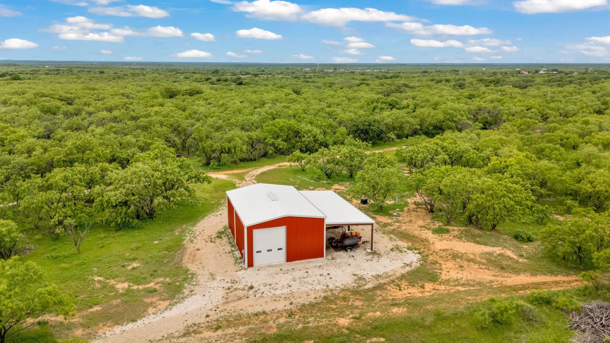 Red metal building with a white roll-up doors and an attached covered lean-to, surrounded by abundant mature trees and natural terrain.