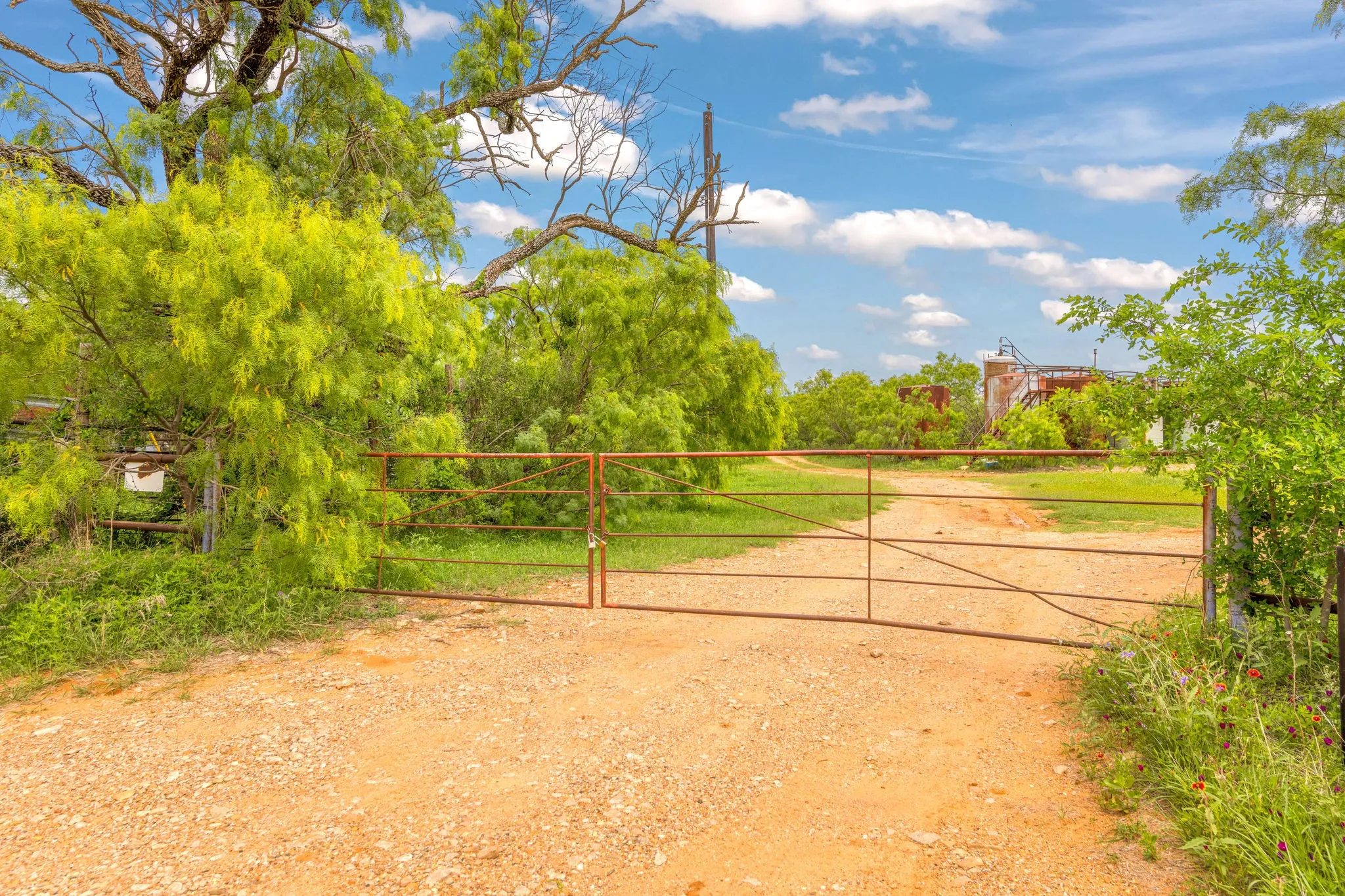 Entrance featuring a double metal gate, surrounded by mature trees and lush vegetation under a clear blue sky, located on CR 133.
