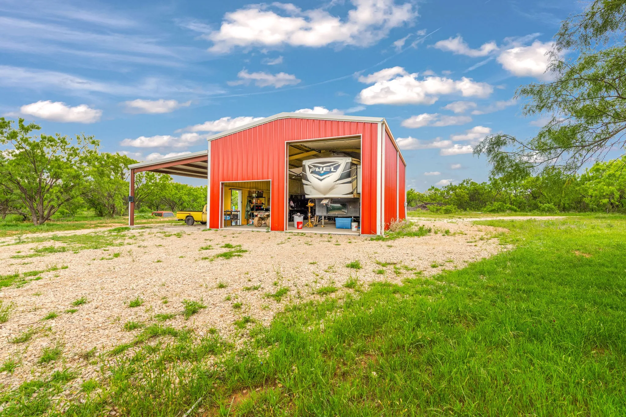 Red metal structure featuring multiple doors, a gravel driveway, and an attached covered carport.