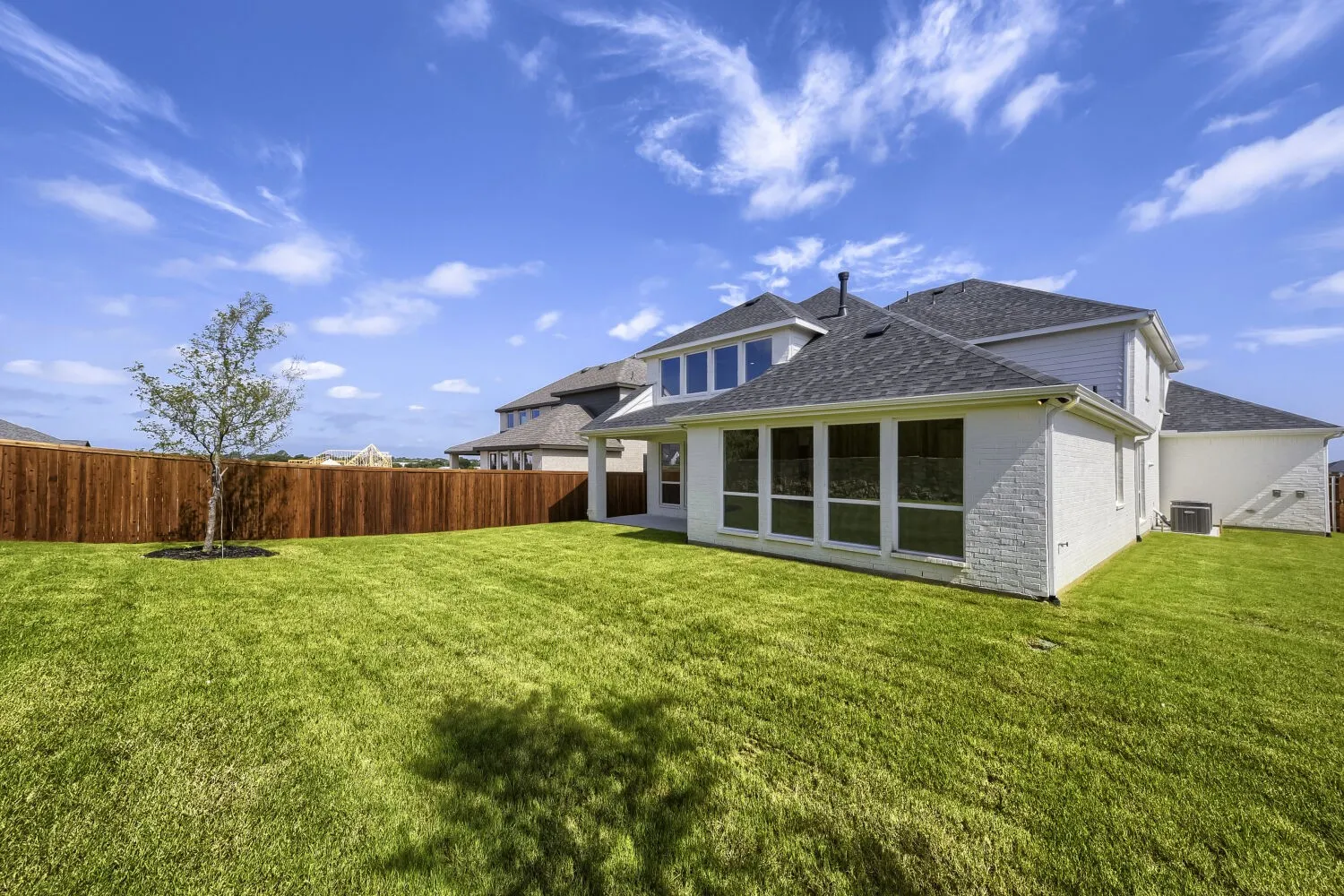 Expansive green lawn bordering a tall privacy fence, featuring a young tree and a two-story residence with white brick and siding, a dark shingle roof, and multiple large windows