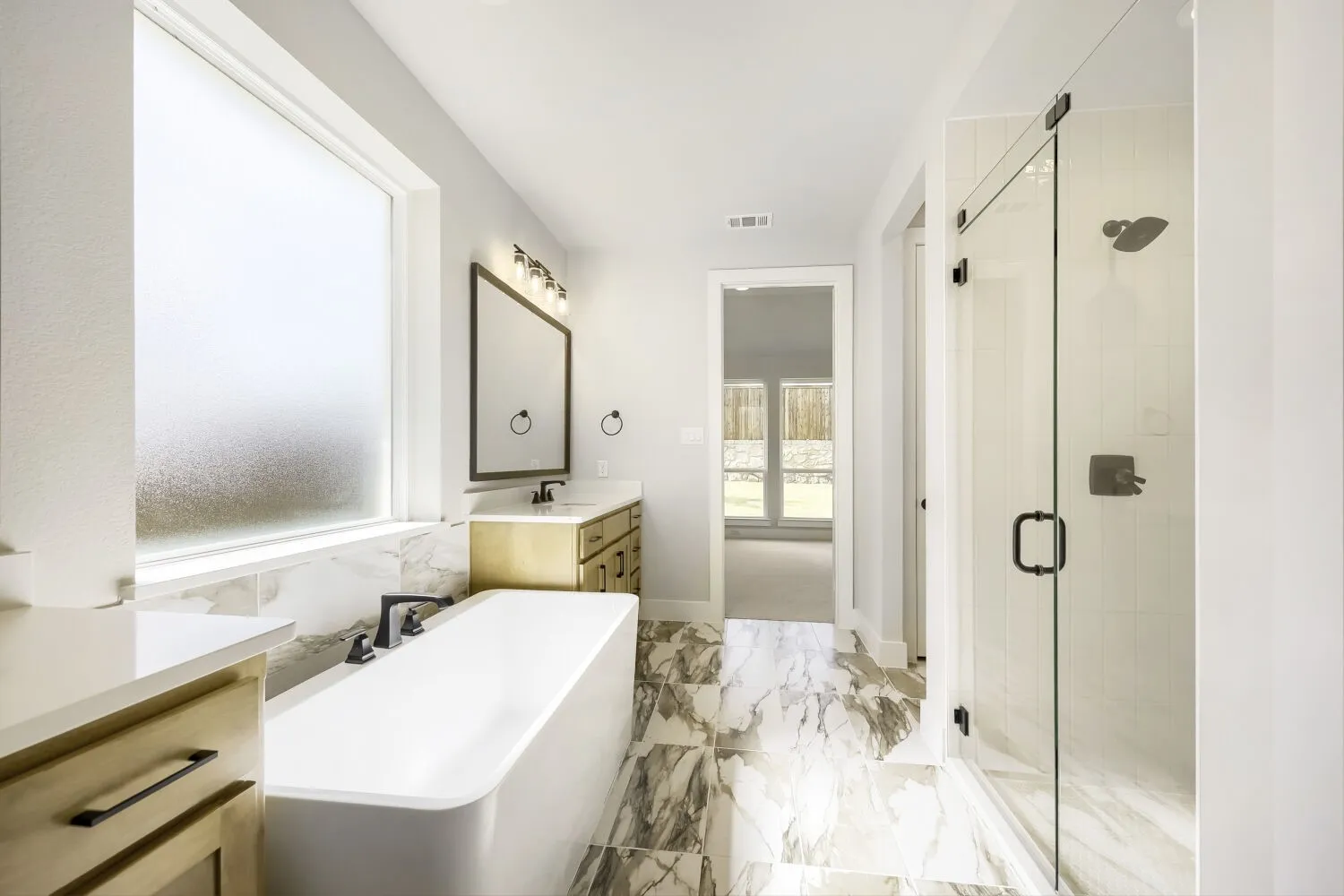 Contemporary bathroom featuring a freestanding soaking tub, a frameless glass shower enclosure, a dual-sink vanity with wood-finish cabinetry, a large rectangular mirror, and marbled tile flooring