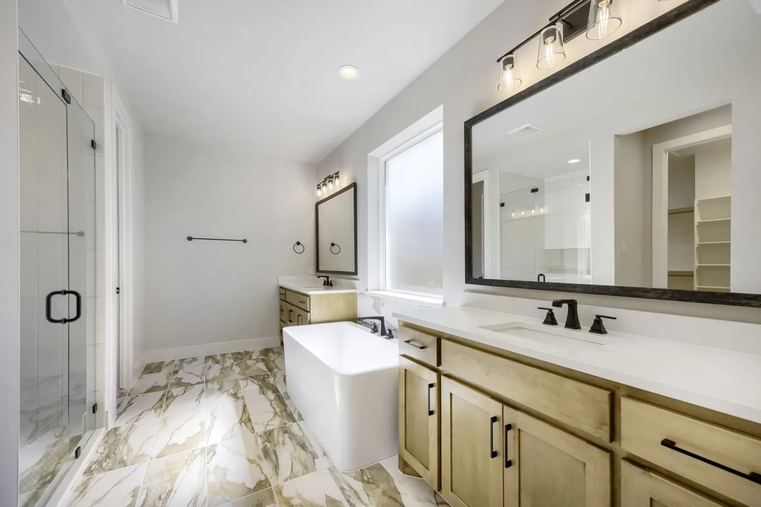 Dual sink vanity with light wood cabinetry and stone-finish countertops, featuring a freestanding soaking tub, a frameless glass shower enclosure, and large-format tile flooring