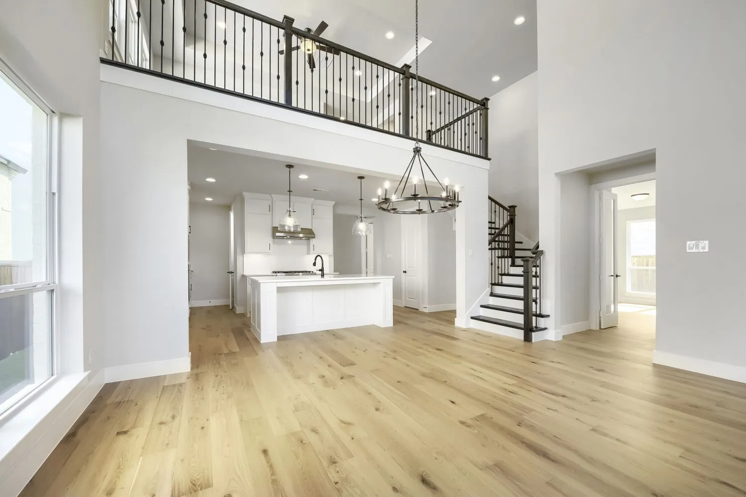 Spacious open-concept interior featuring light wood-finish flooring, a two-story ceiling, a contemporary chandelier, a white island kitchen with pendant lighting, and a prominent staircase with dark iron balusters