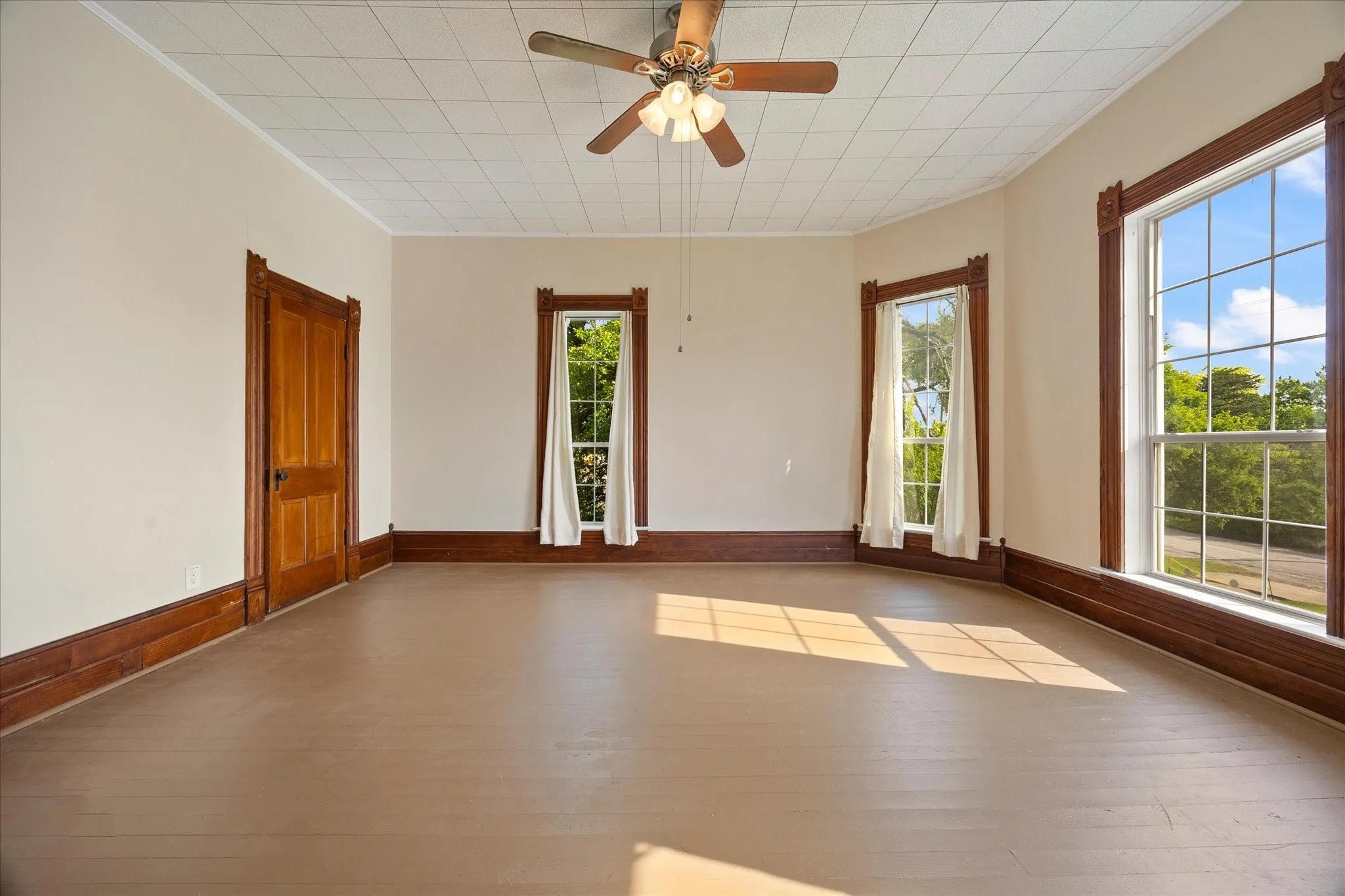 Spacious room featuring light-toned flooring, tall windows with decorative wood trim, and a ceiling fan