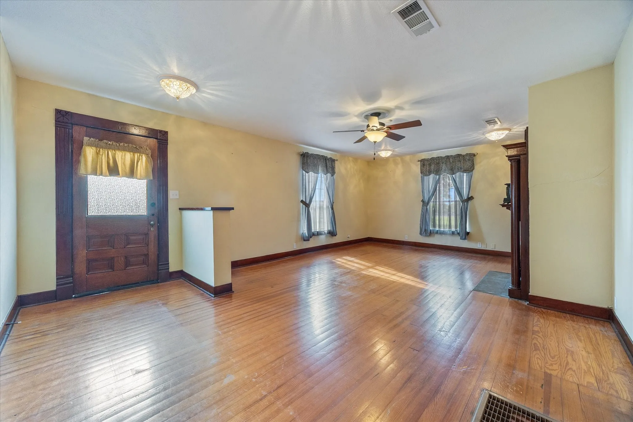 Spacious room featuring wood-finish flooring, multiple windows with drapes, a ceiling fan with integrated lighting, and additional ceiling-mounted light fixtures