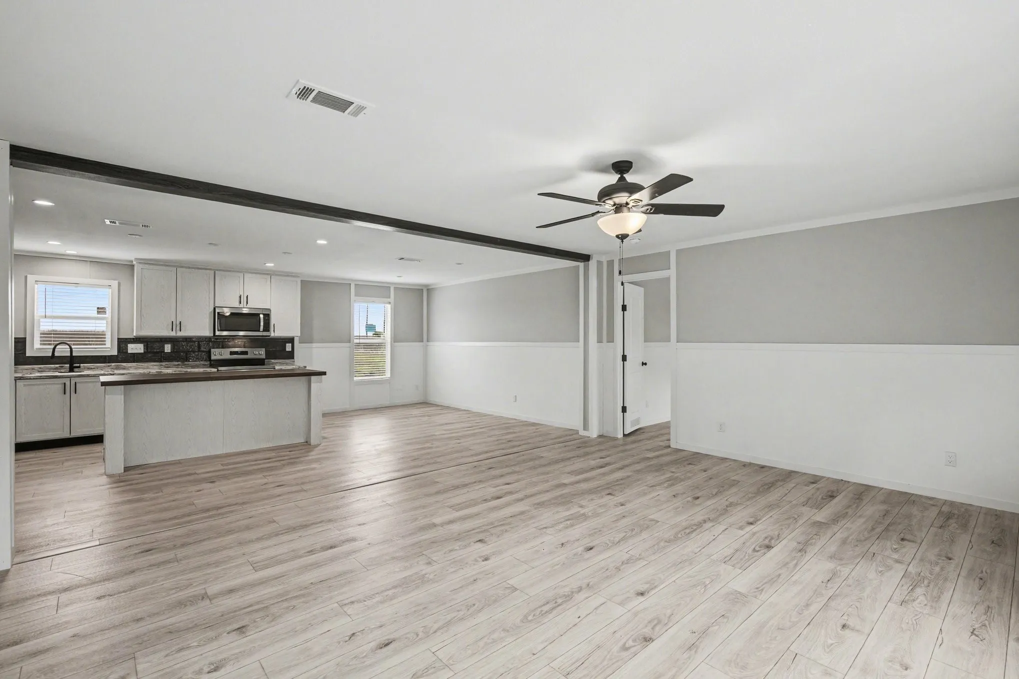 Spacious open-concept room featuring wood-look flooring, a contemporary ceiling fan with integrated lighting, and a kitchen area with light-toned cabinetry, stainless steel appliances, and dark countertops