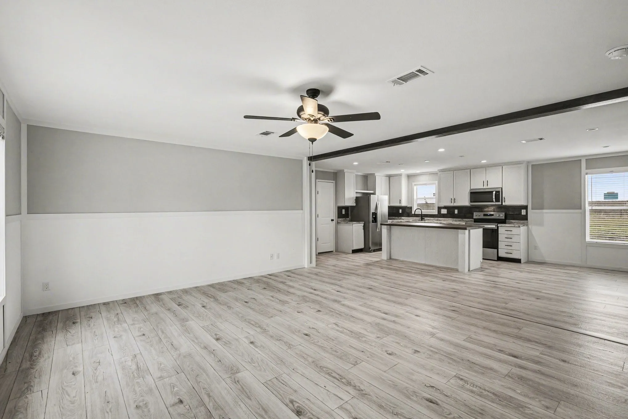 Spacious open-concept living area featuring wood-look flooring, a ceiling fan with integrated lighting, and a kitchen with white cabinetry, dark countertops, and stainless steel appliances