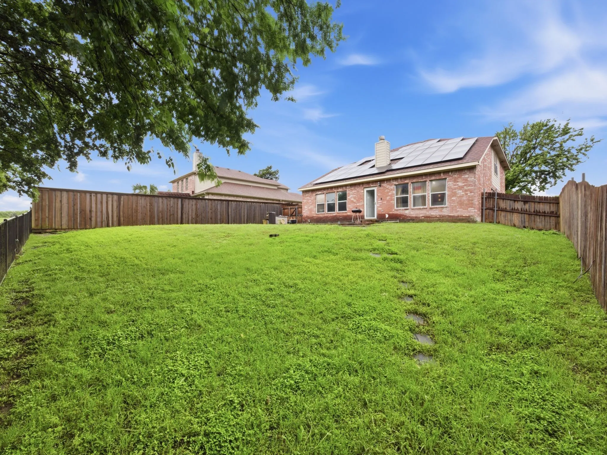 Expansive grassy backyard featuring a brick exterior home with multiple windows, a rear entry door, and a brown shingle roof with solar panels