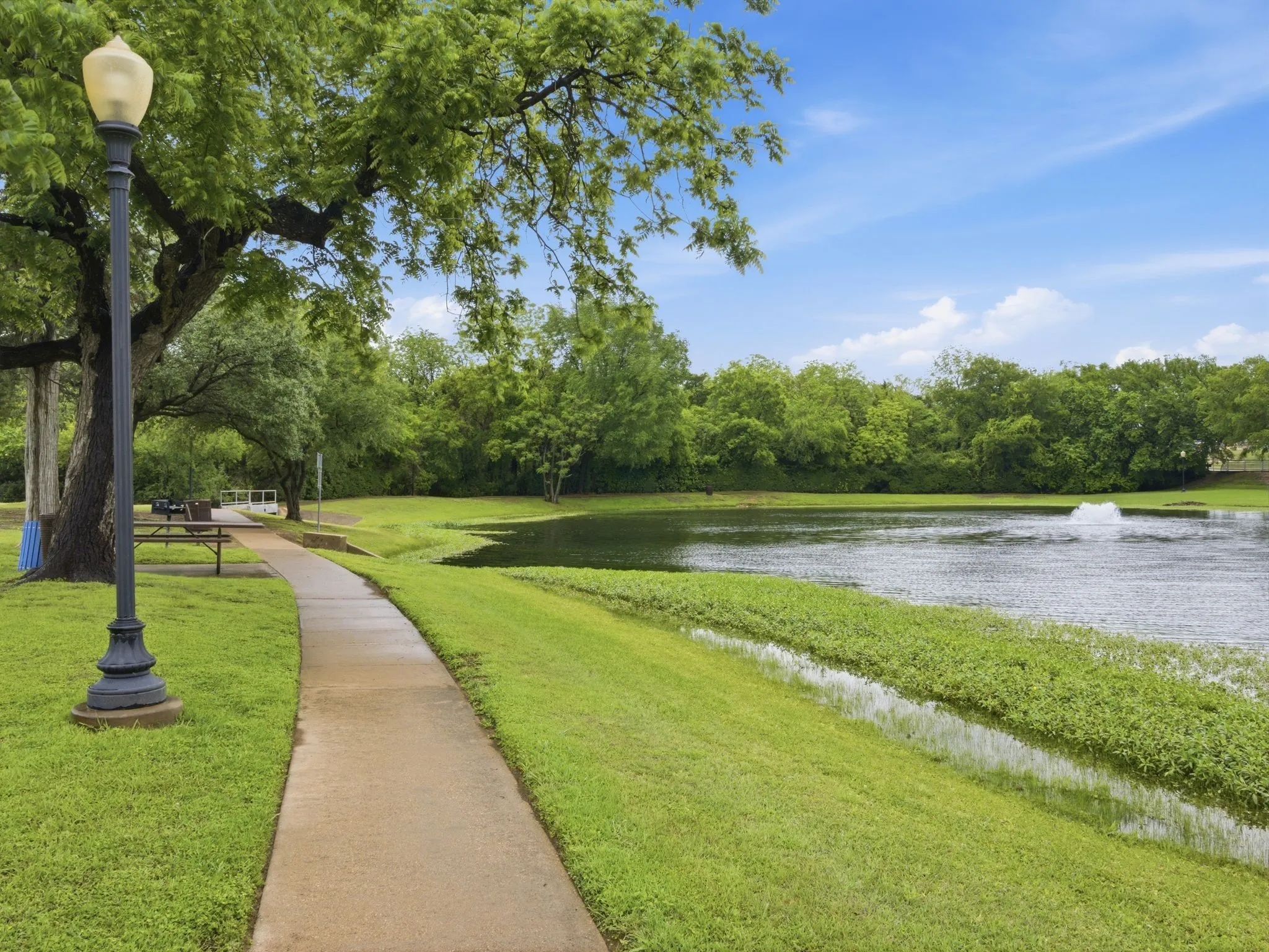 Landscaped grounds featuring a paved pathway, mature trees, a tranquil pond with a fountain, and abundant green space