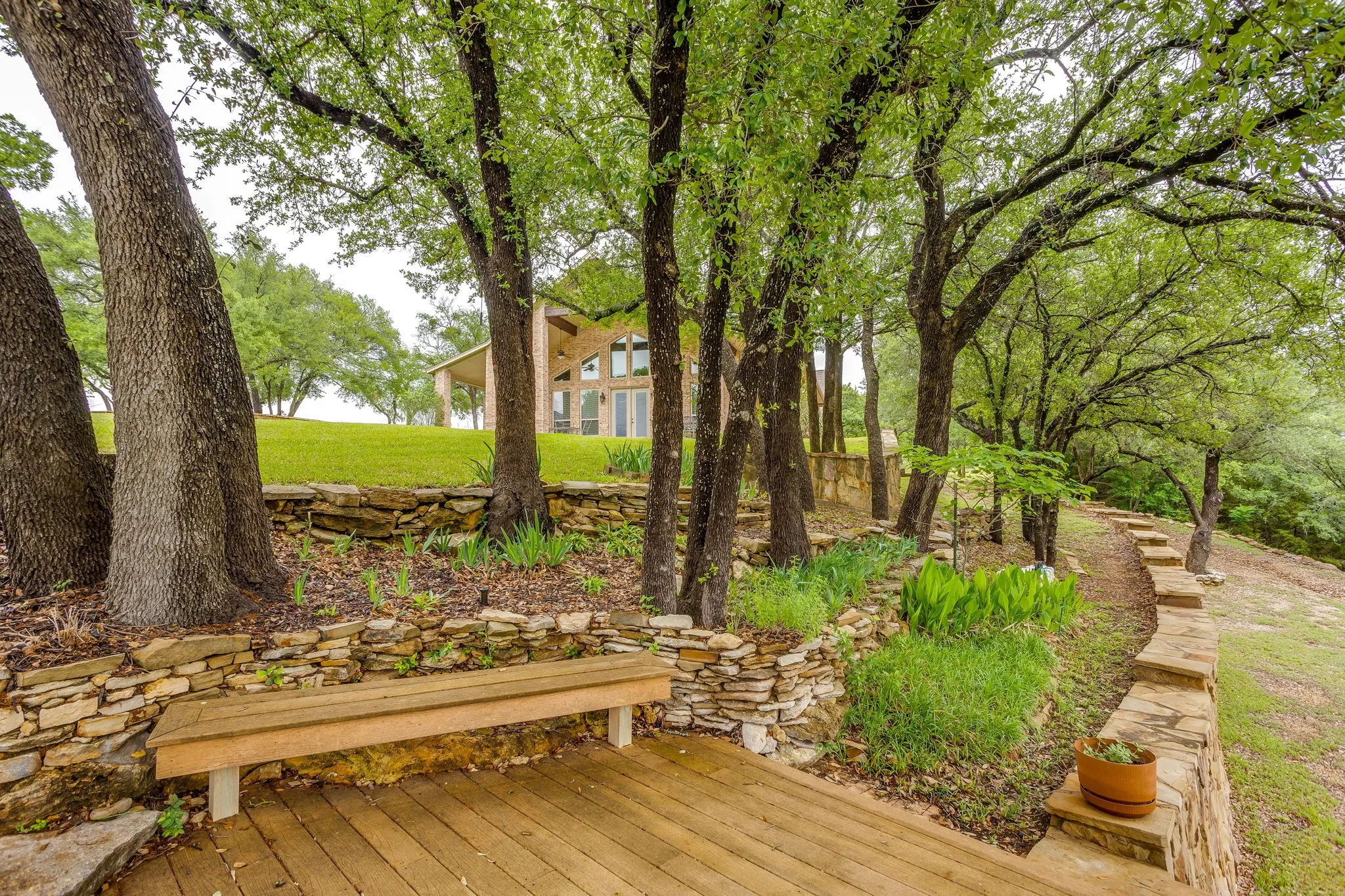 Tiered landscaping featuring a wood-finish deck with built-in bench seating, stacked stone retaining walls, and mature trees