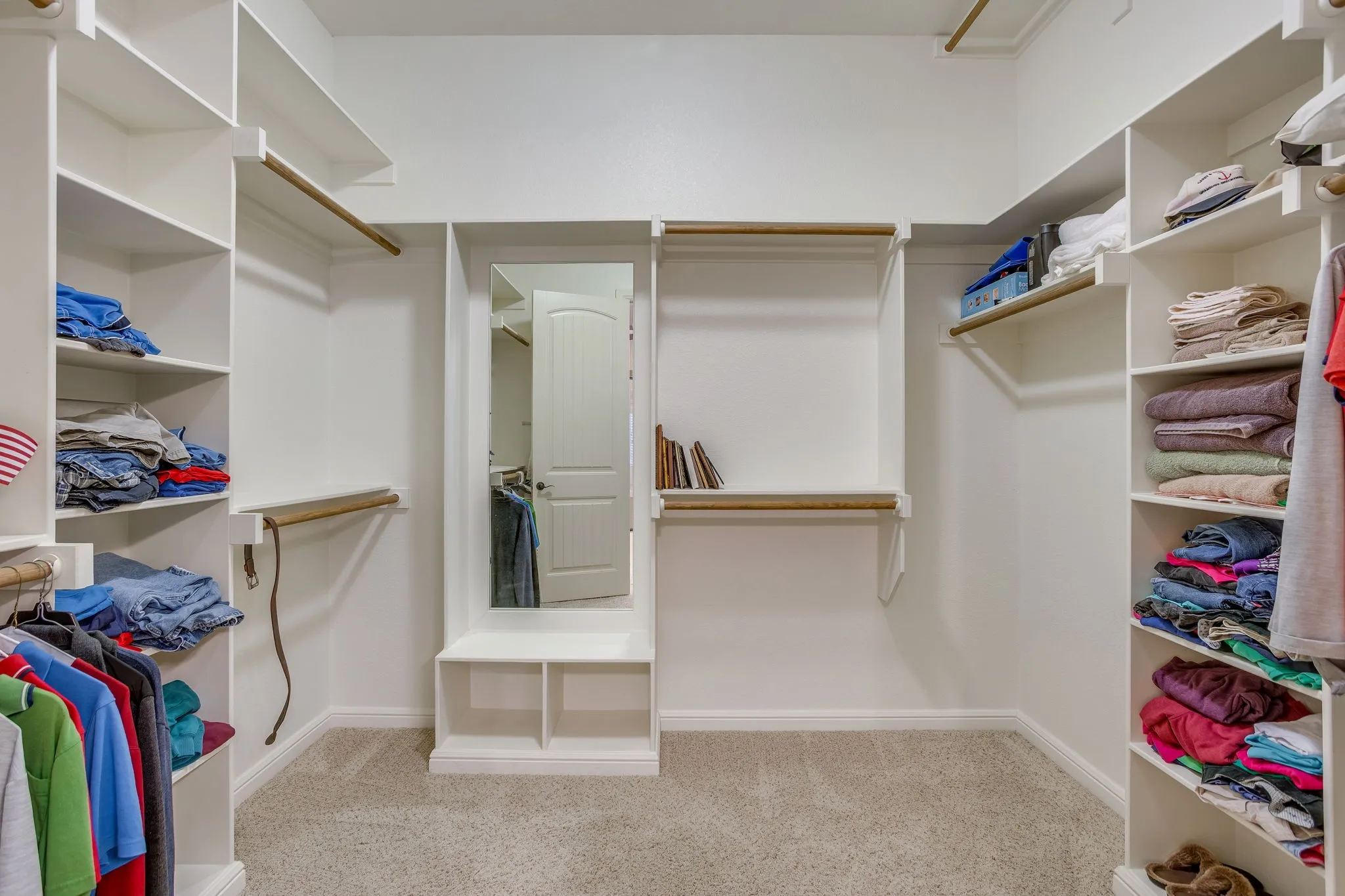 Spacious walk-in primary closet featuring built-in shelving, multiple hanging rods, integrated full-length mirror, and neutral carpet flooring