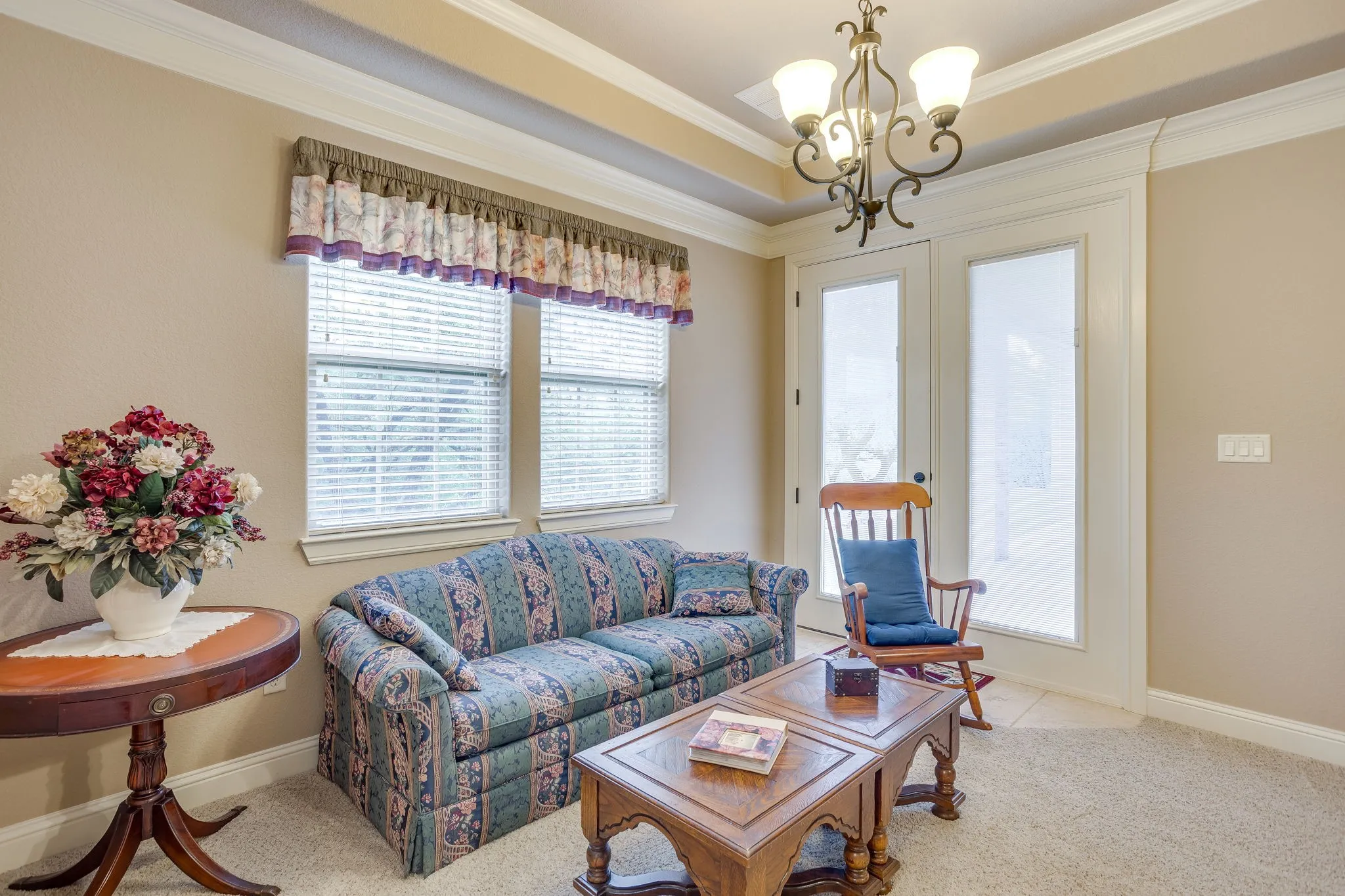 Seating area in primary bedroom with private access to outdoor covered porch, featuring a double window with blinds, a tray ceiling with crown molding, and an ornate chandelier