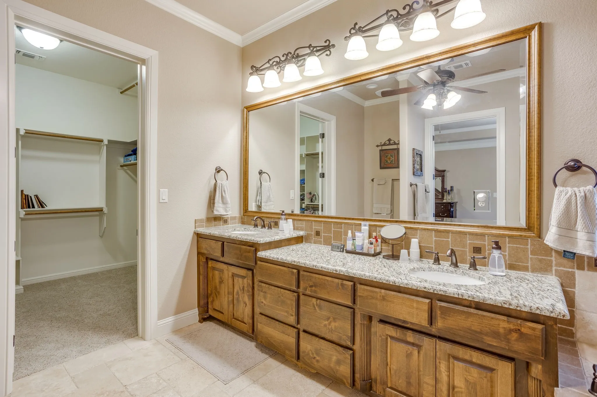 Primary bathroom featuring a dual vanity with light-toned granite countertops, wood-finish cabinetry, and a large framed mirror with ornate lighting