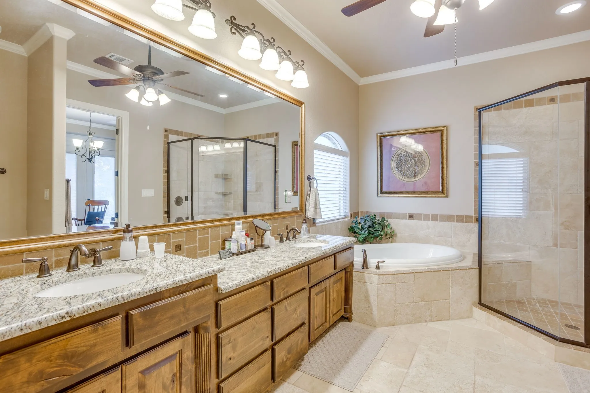 Primary bathroom featuring a dual vanity with granite countertops and wood cabinetry, an oval soaking tub with tiled surround, a walk-in shower with glass enclosure, arched window with horizontal blinds, and tile flooring