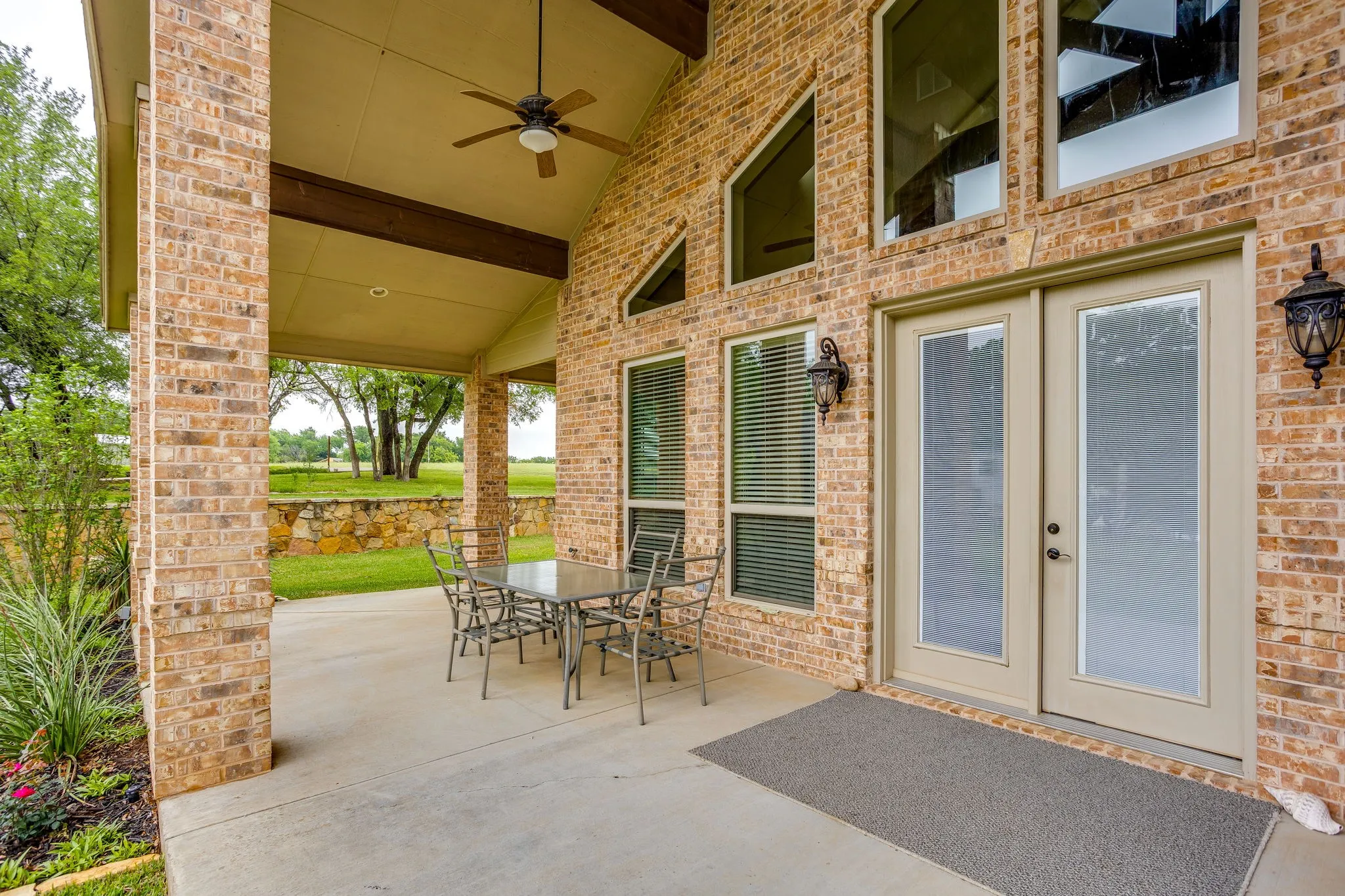 Expansive covered patio with a vaulted ceiling and ceiling fan, featuring brick columns and a stone retaining wall