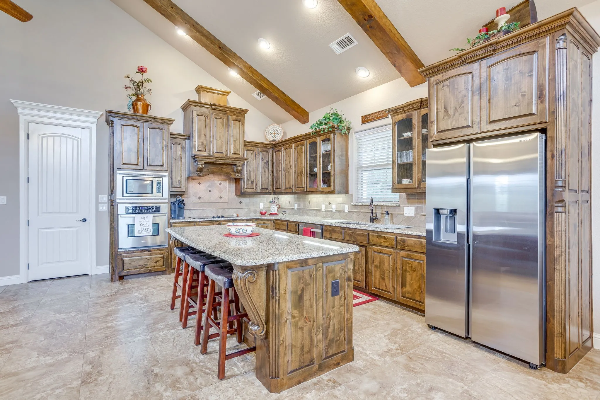 Spacious kitchen featuring vaulted ceilings with exposed wood beams, custom wood cabinetry, a large kitchen island with counter seating, stainless steel appliances, and tile flooring