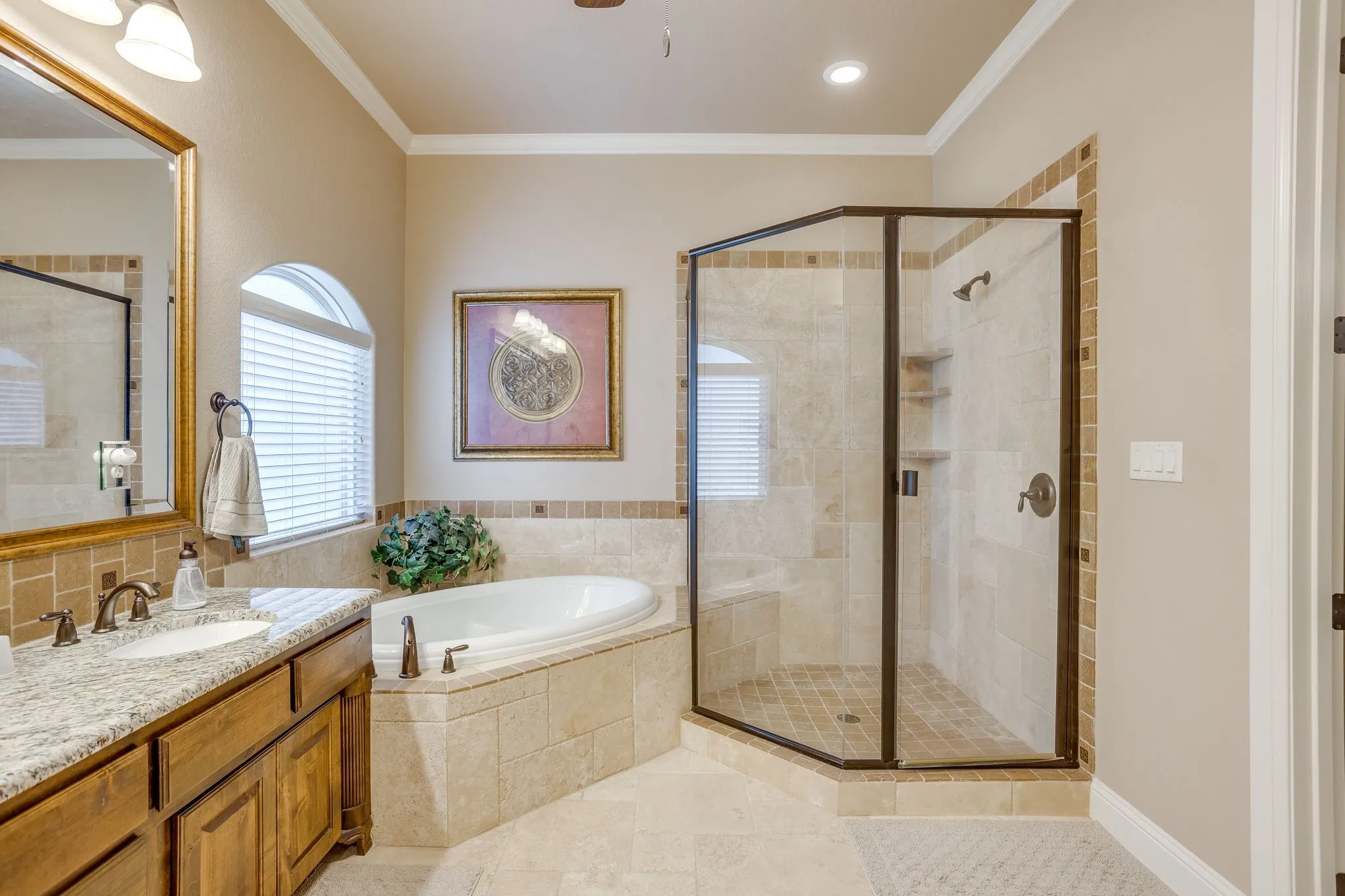 Primary bathroom featuring a corner soaking tub, a separate glass-enclosed shower with tiled walls and built-in shelving, a dual-sink vanity with a granite-style countertop, and a large arched window with horizontal blinds