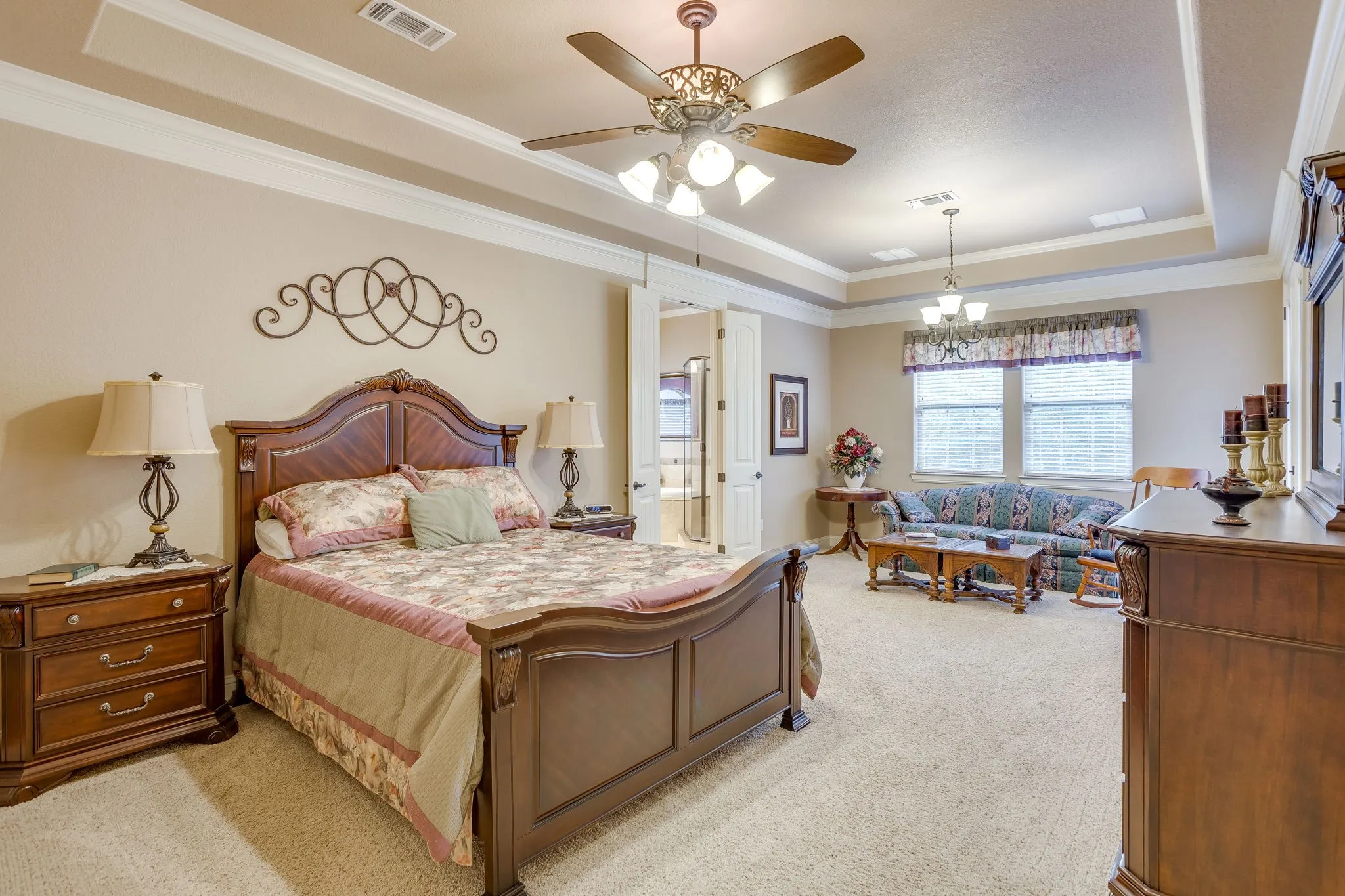 Spacious carpeted primary bedroom room featuring a tray ceiling with crown molding, two windows with blinds, and a ceiling fan with integrated lighting