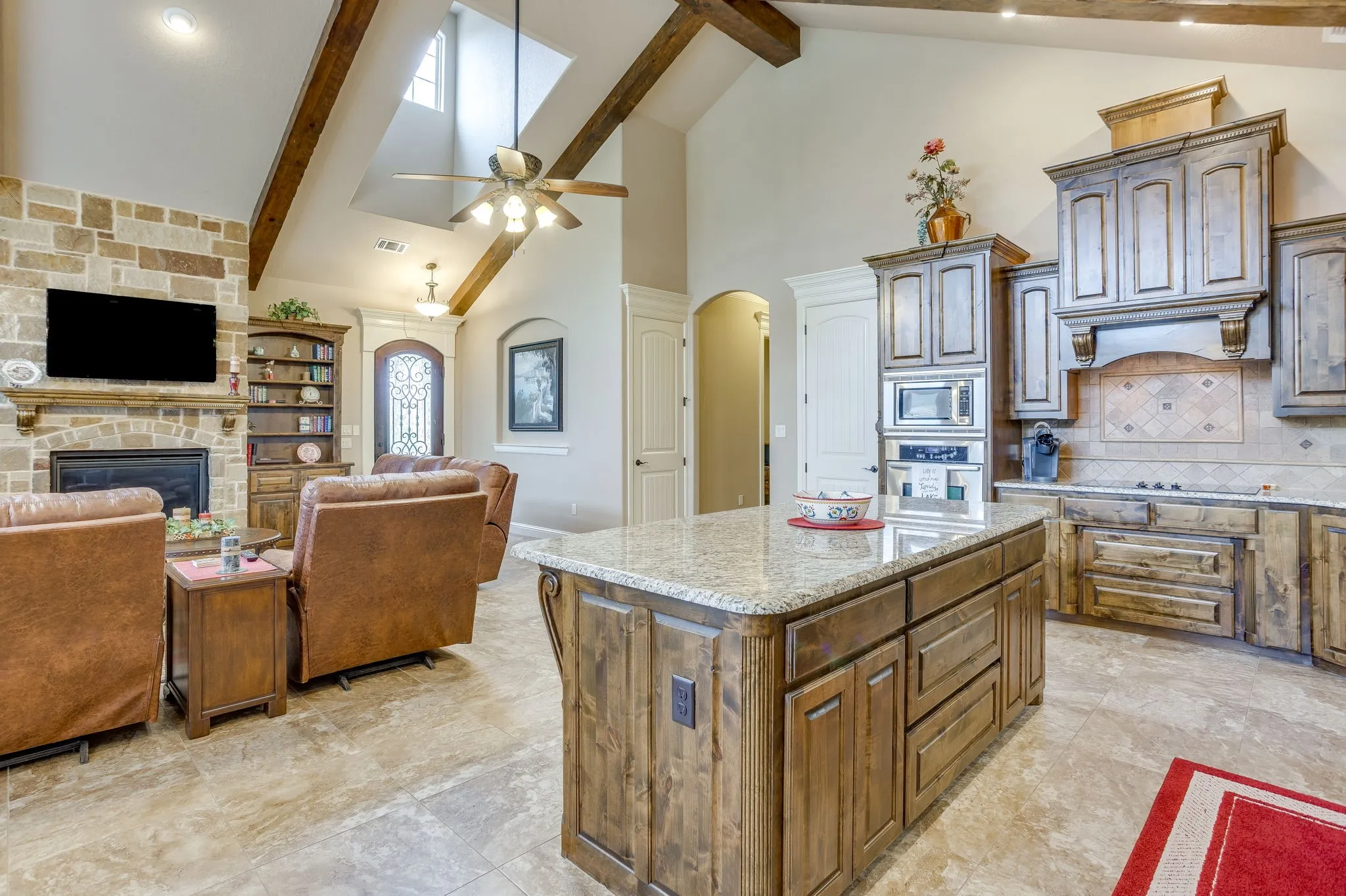 Open-concept living space with a kitchen island featuring a granite countertop, a stone-clad fireplace, exposed wooden ceiling beams, and vaulted ceilings