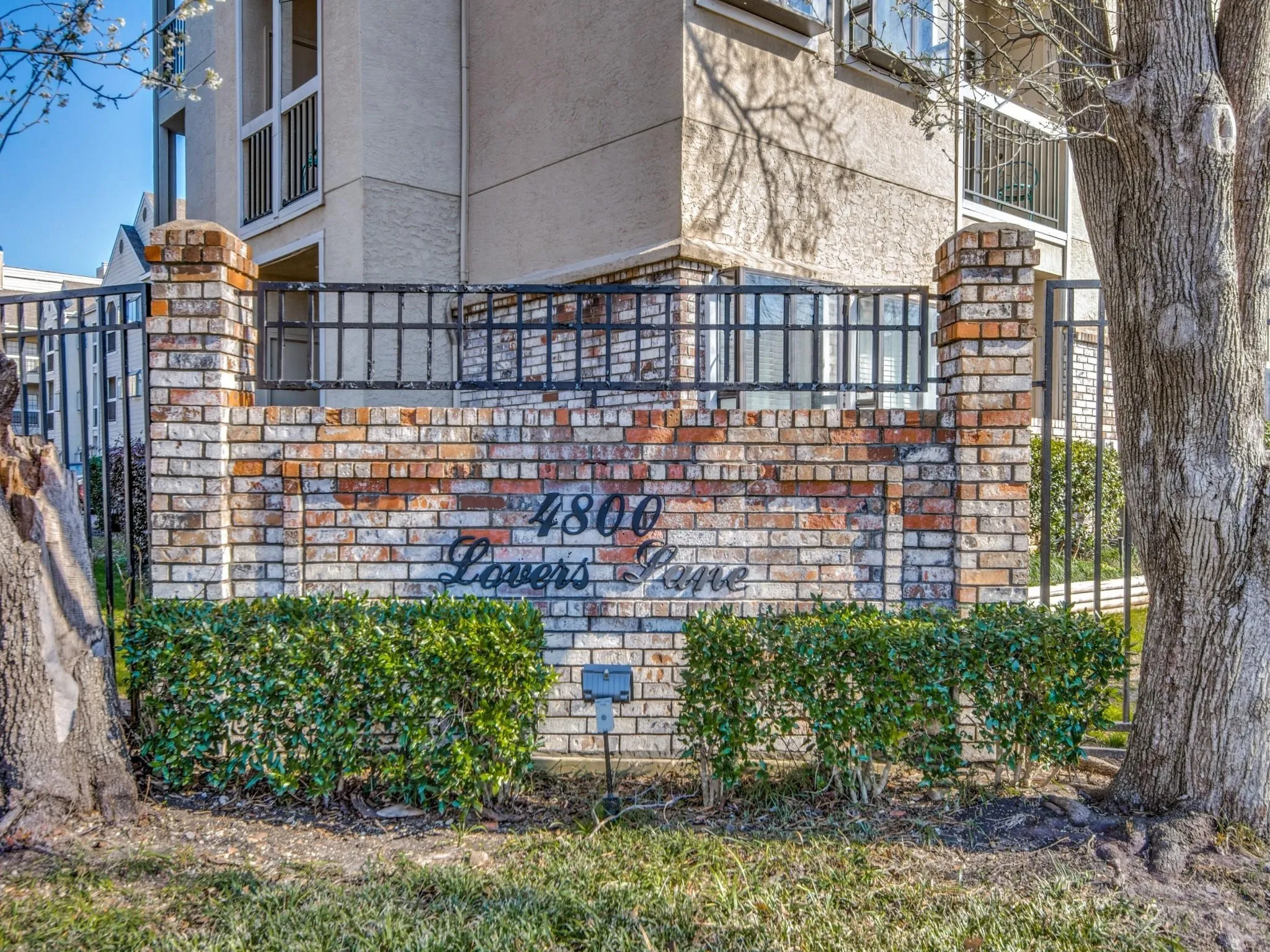 Brick facade entrance signage featuring wrought iron fencing and manicured shrubbery