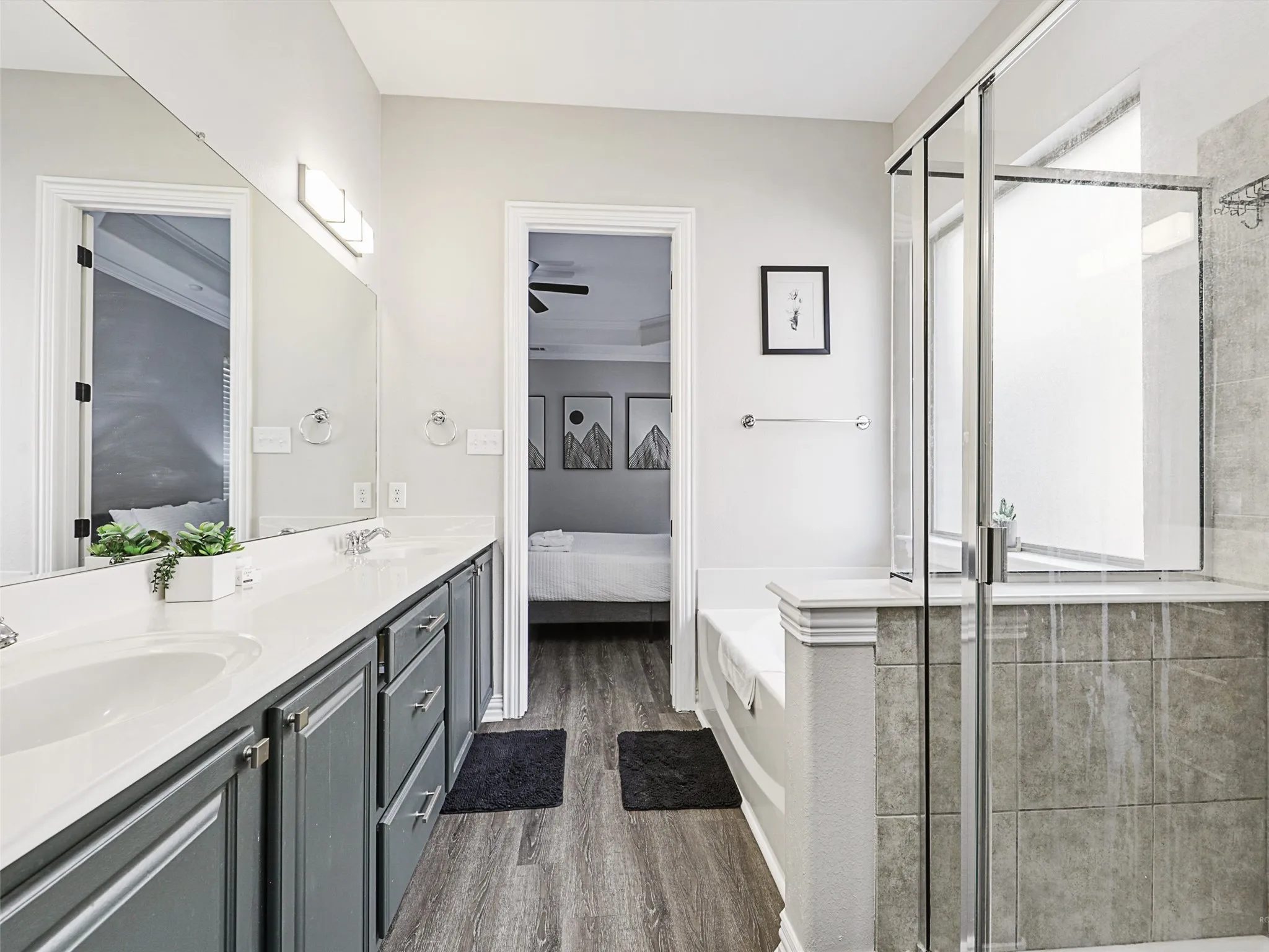 Bathroom featuring a double vanity with grey cabinetry, a spacious built-in tub, a glass-enclosed shower, wood-finish flooring, and a large wall-mounted mirror
