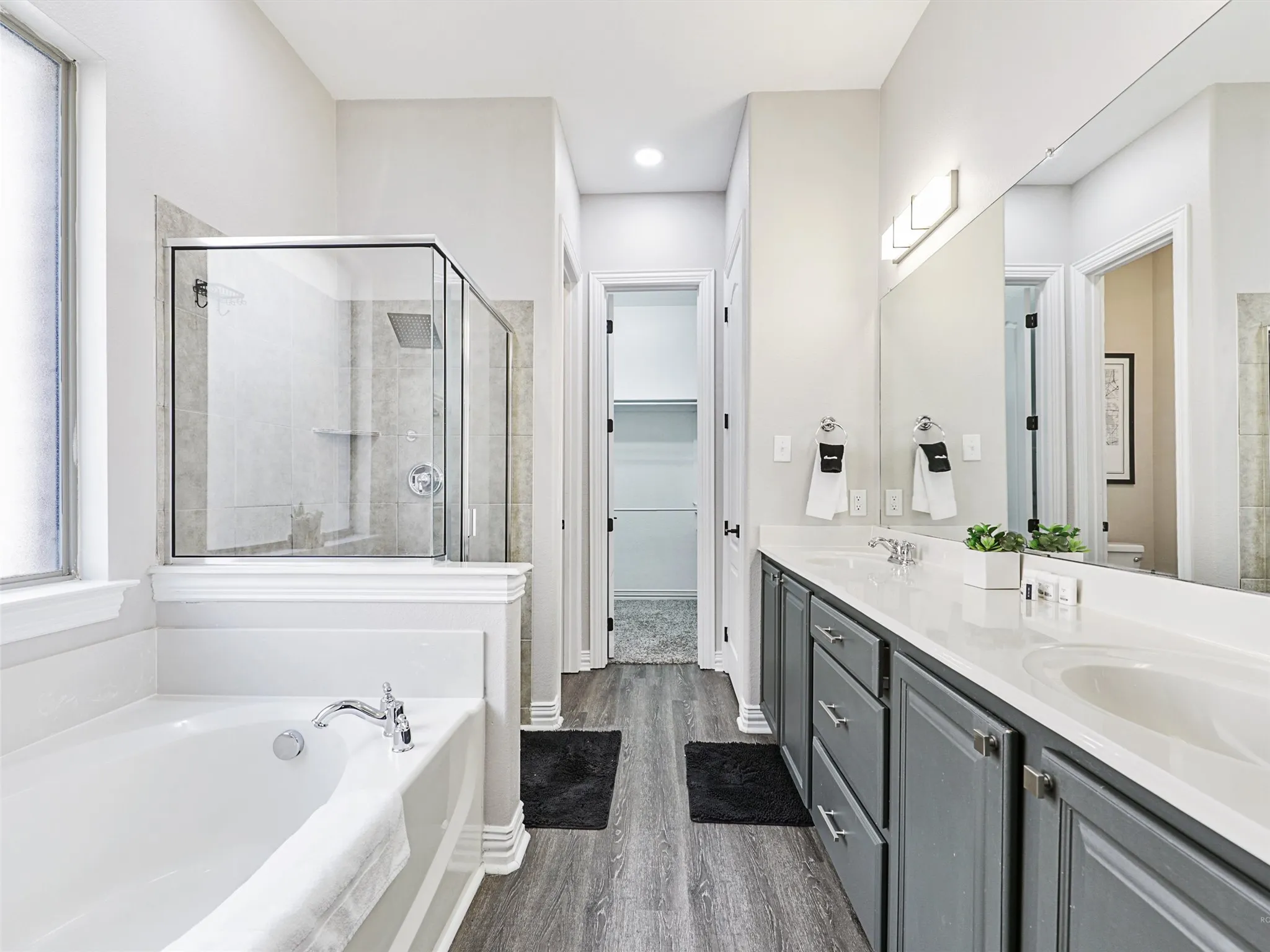 Spacious bathroom featuring a built-in soaking tub, glass-enclosed shower, dual vanity with undermount sinks, wood-finish flooring, and a walk-in closet