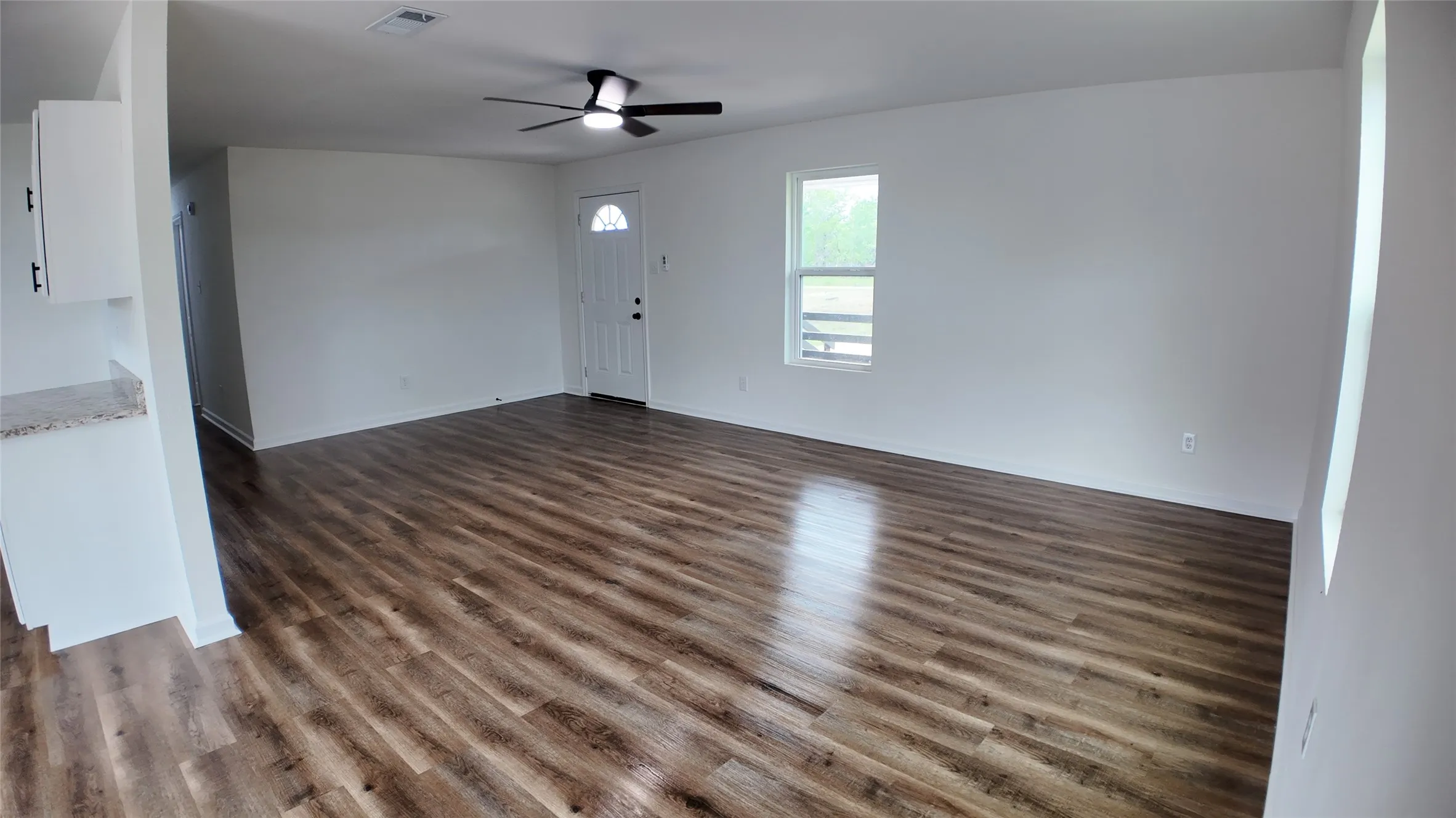 Open concept living area featuring wood-finish flooring, white walls, a ceiling fan with integrated lighting, a front entry door with an arched glass insert, and a window providing exterior views