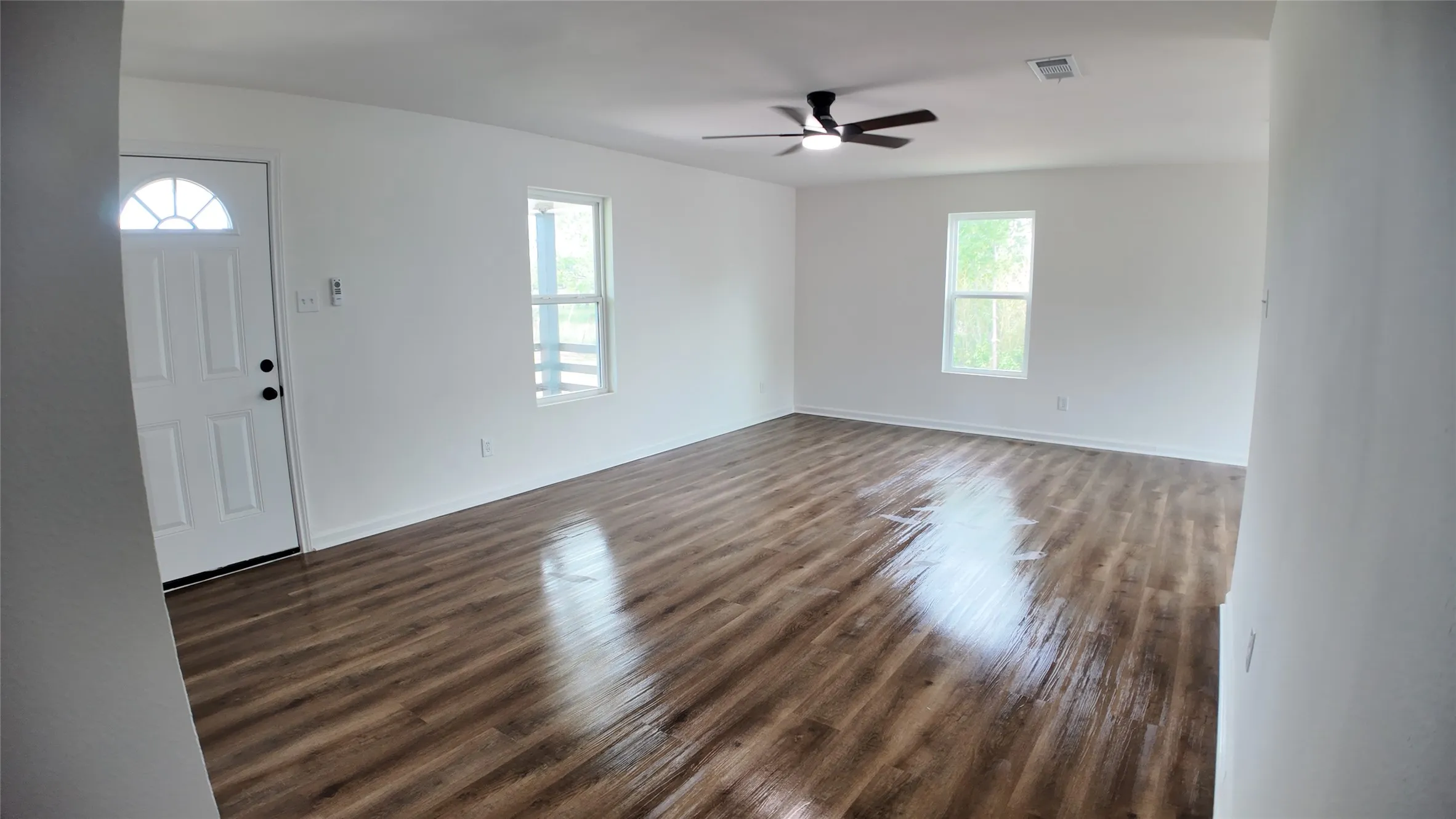 Spacious room featuring wood-finish flooring, white walls, multiple windows, a ceiling fan, and a front entry door with an arched glass insert