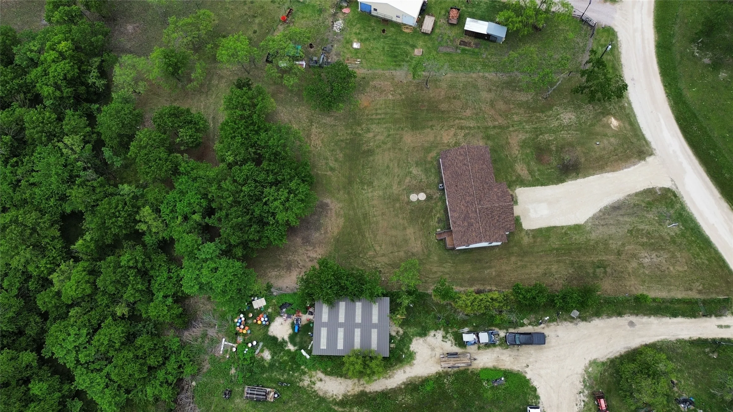Aerial property view featuring a gravel driveway, a single-story residence with a brown shingle roof, a secondary structure with a metal roof, and extensive natural tree coverage