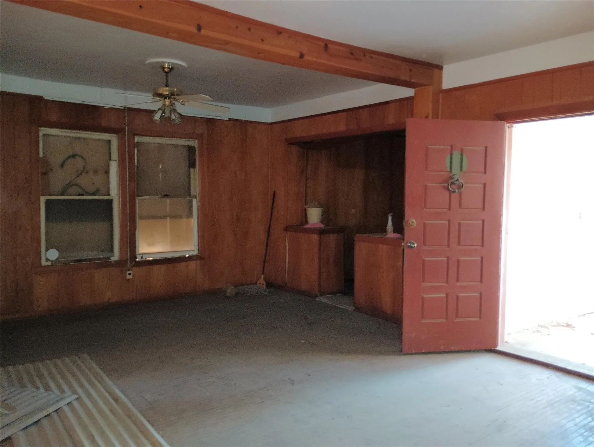 Spacious room featuring wood paneling, exposed wood ceiling beams, two windows, built-in wooden cabinetry, and a red paneled door