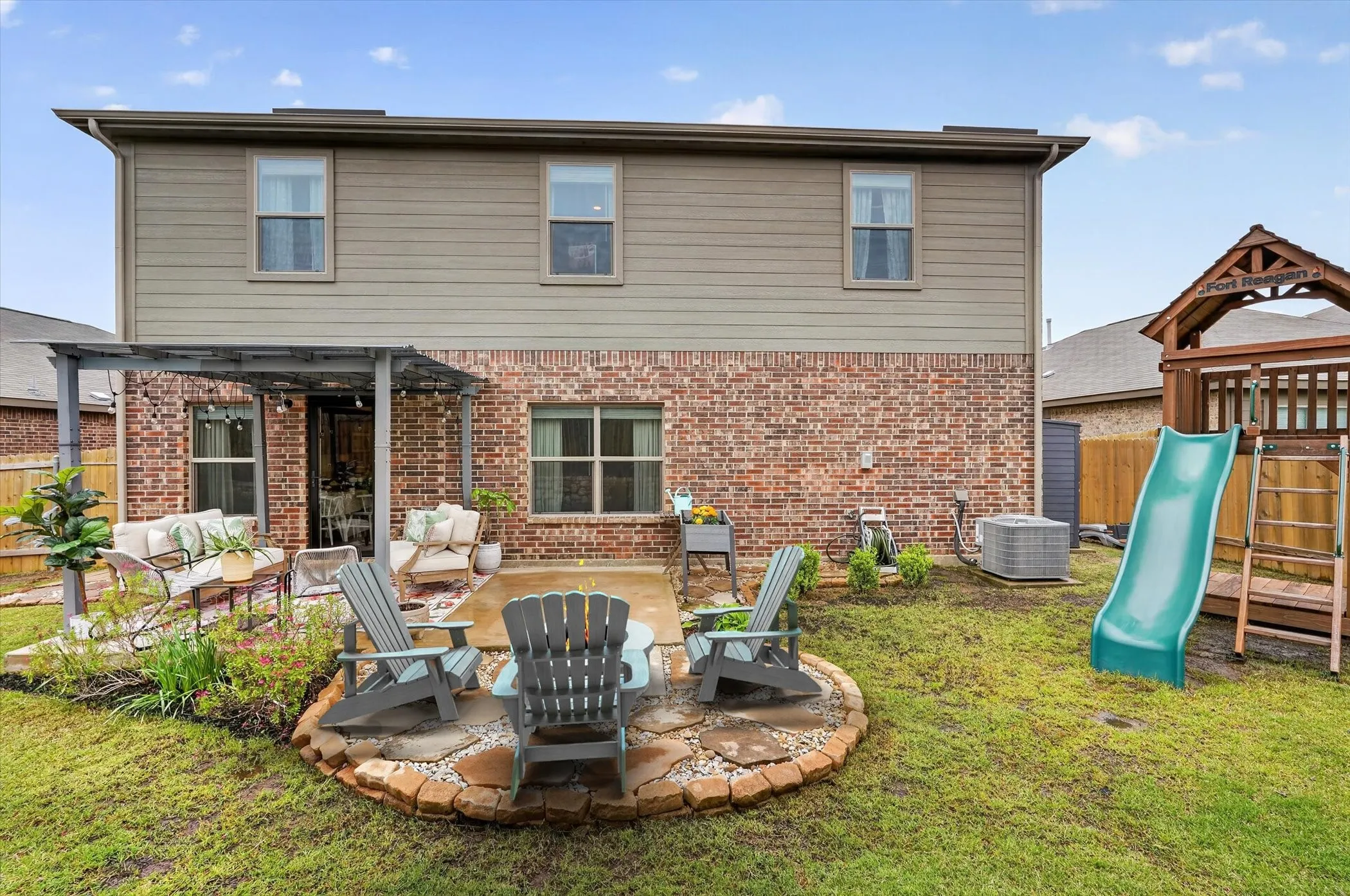 Virtual staged Firepit.Expansive backyard featuring a brick and siding exterior, a covered patio with string lights, a stone-bordered fire pit area, and a green lawn