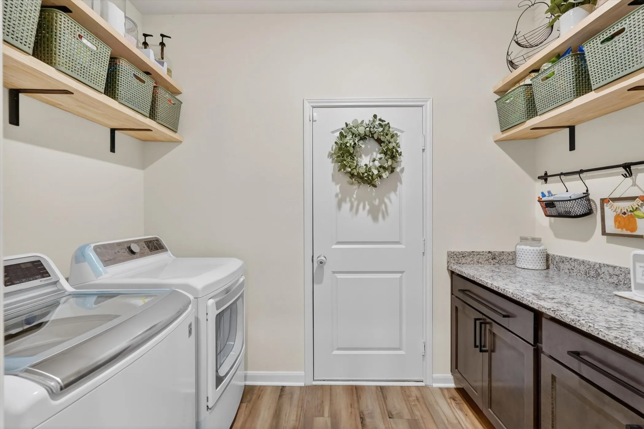 Laundry area featuring wood-finish flooring, light-toned walls, floating wood shelving with black brackets, dark lower cabinetry, and a light-toned countertop