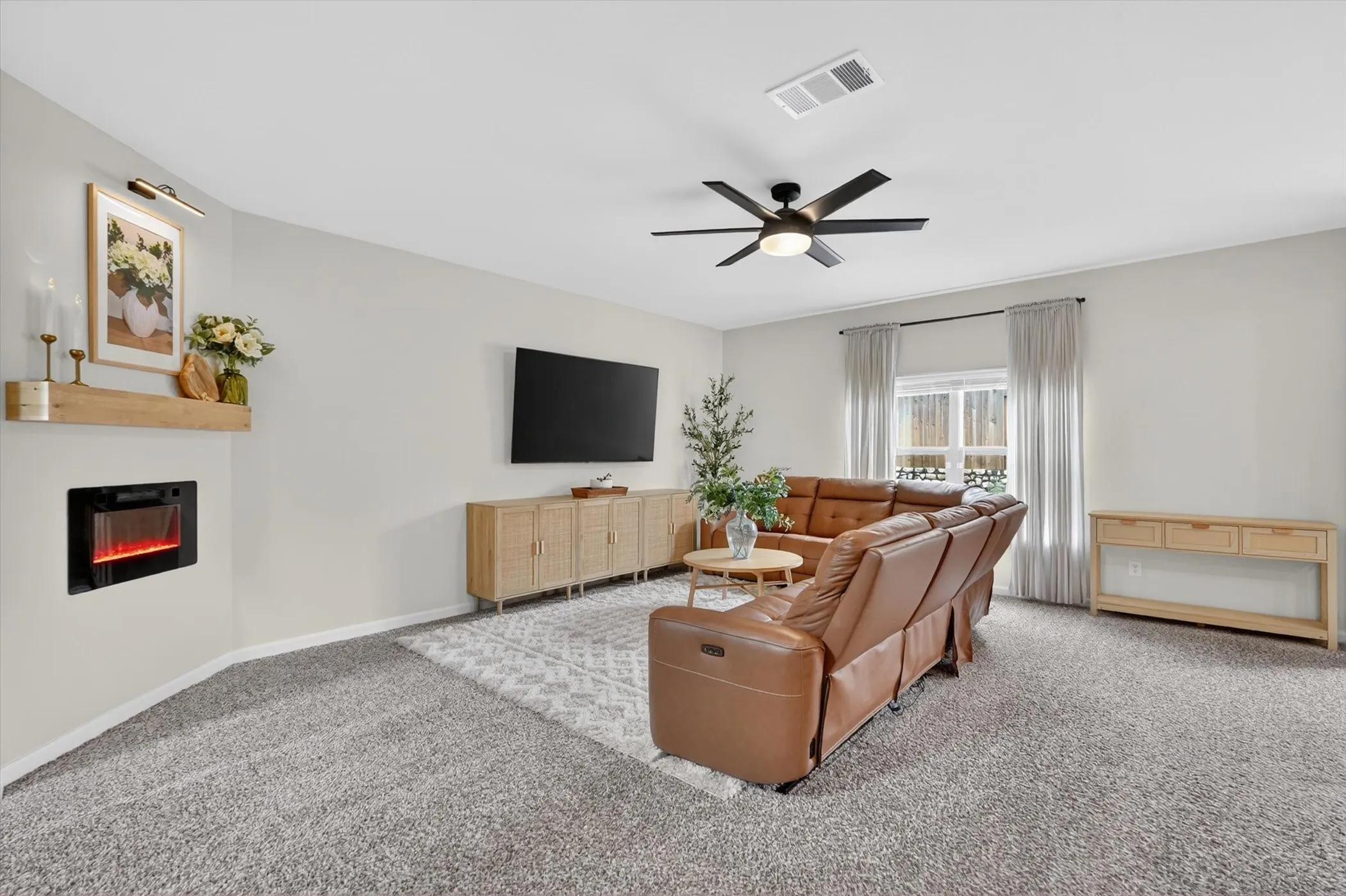 Living area featuring a corner-mounted electric fireplace, a wood mantle, a ceiling fan with integrated lighting, window treatments, and carpet flooring
