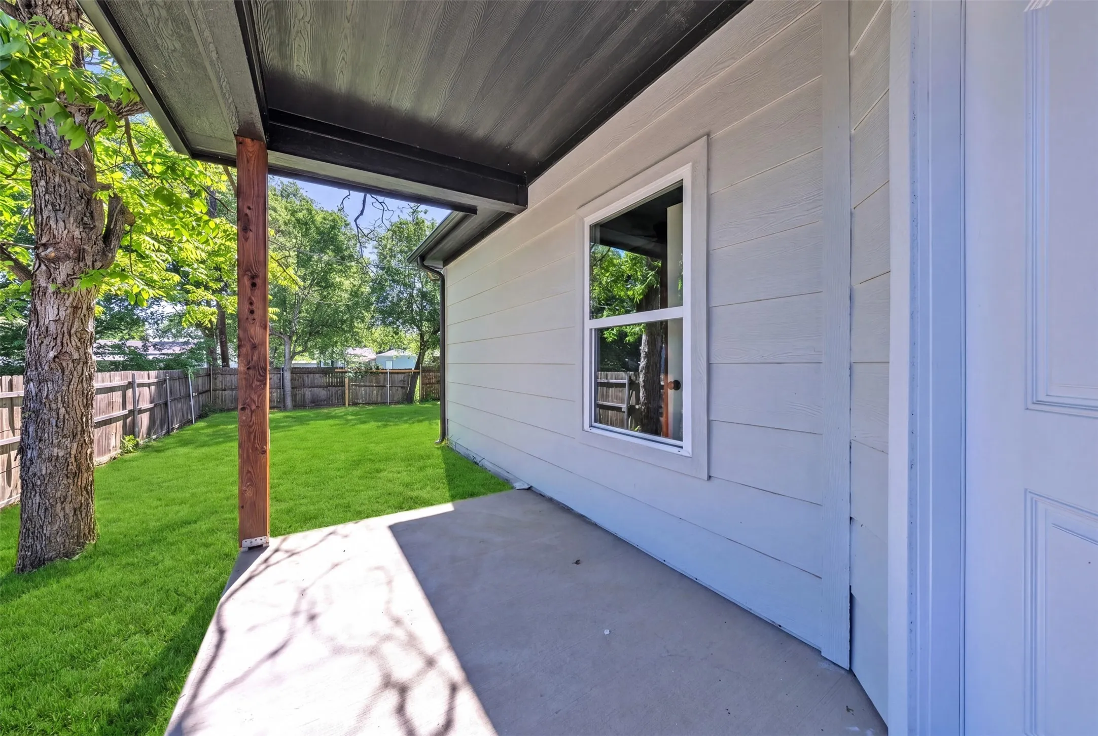 Covered concrete patio with a single wood support beam, wood plank siding, and a white-framed window