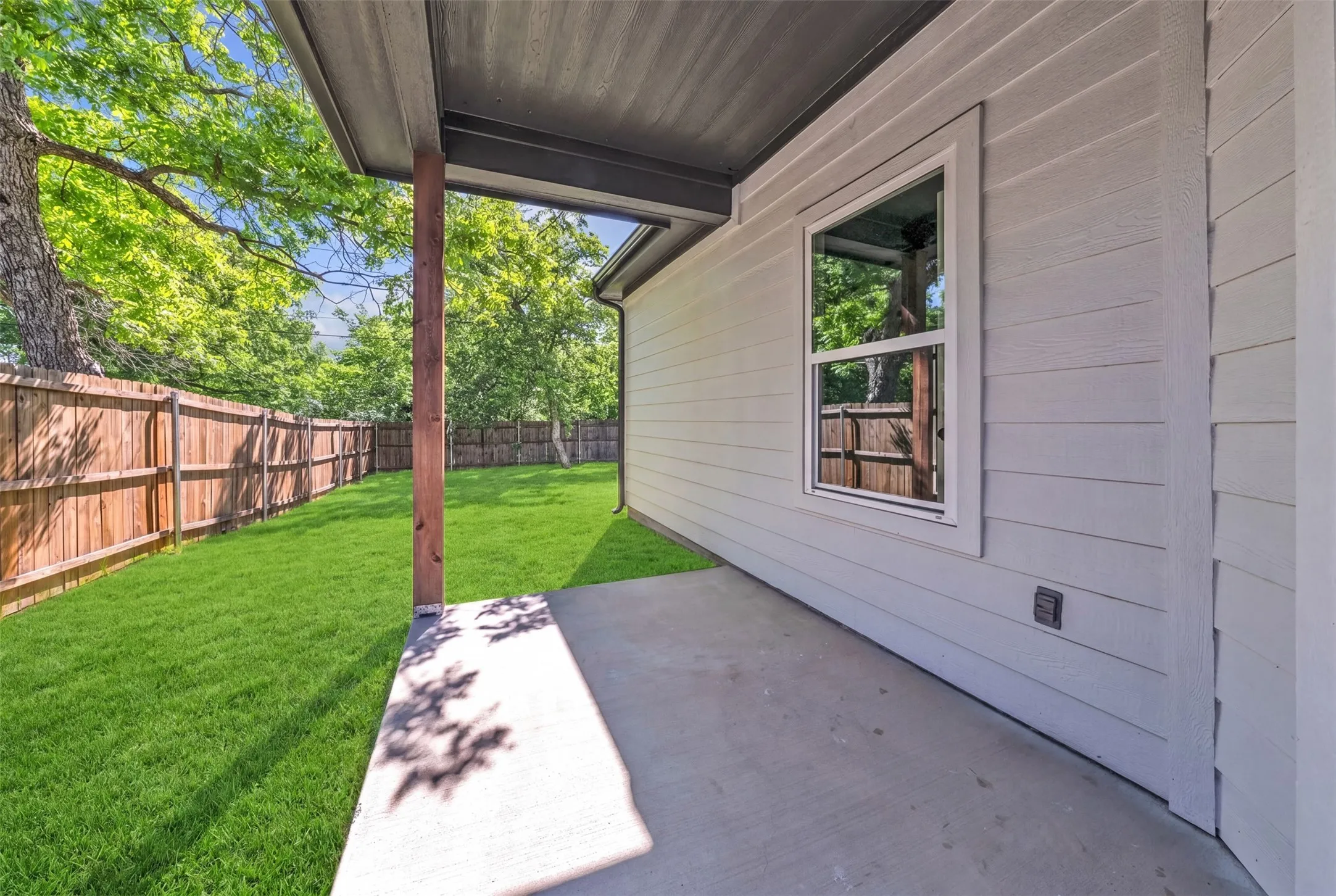 Covered concrete patio extending to a fenced backyard with lush green lawn and mature trees