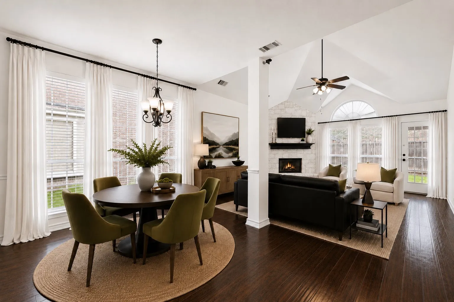 Vaulted ceiling with a ceiling fan, fireplace with a stone-finish facade, wood-finish flooring, multiple windows with blinds, and an exterior door; virtually staged