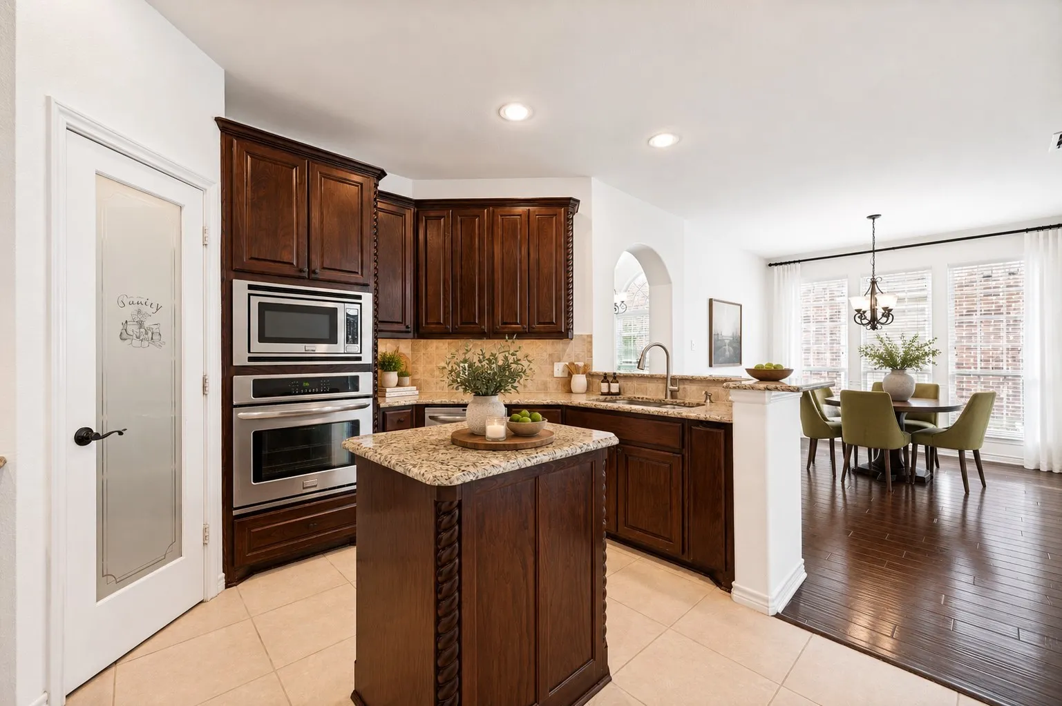 Kitchen featuring warm wood cabinetry, stainless steel double ovens, a central island with a granite countertop, and tile flooring; virtually staged.