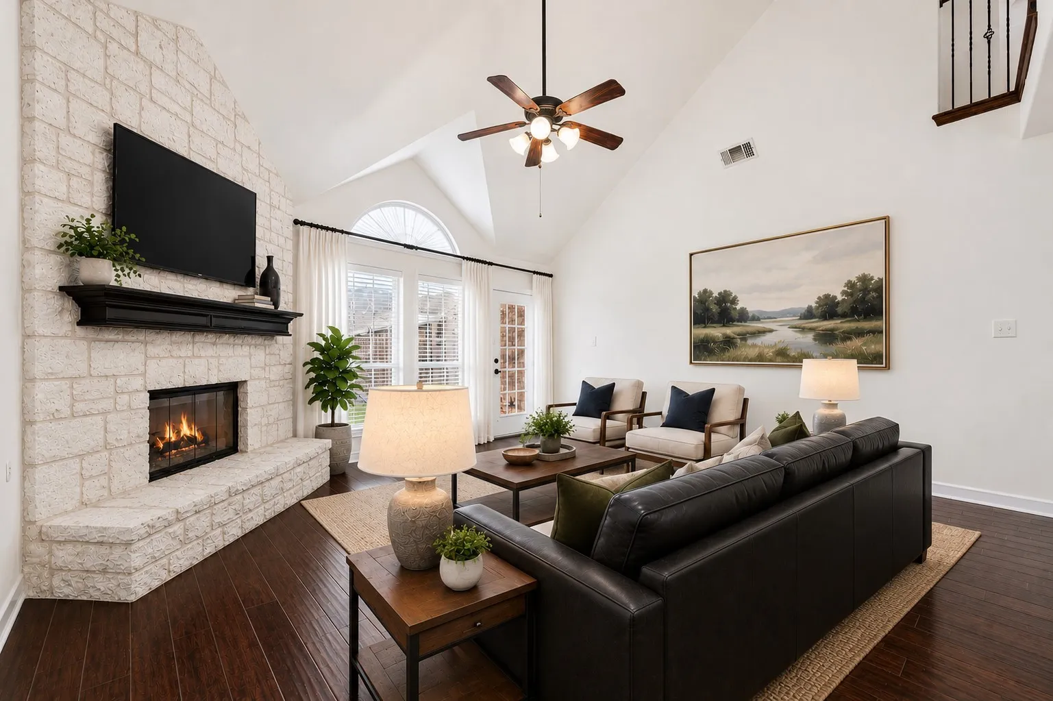 Stone-faced fireplace with a dark wood mantel, high vaulted ceiling, wood-finish flooring, ceiling fan, and French doors with an arched transom window; virtually staged