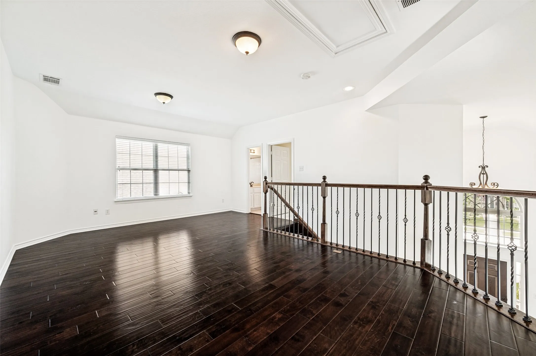 Spacious loft featuring dark wood-finish flooring, a large window with horizontal blinds, and a decorative wrought iron and wood railing overlooking the entry