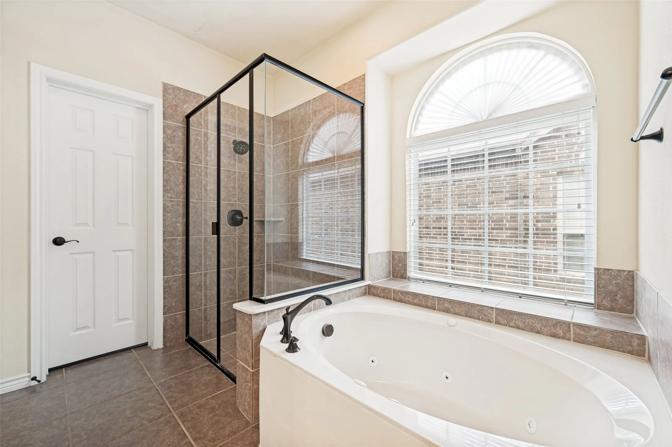 Bathroom featuring a jetted soaking tub, separate glass-enclosed shower with tile surround, and a large arched window with blinds
