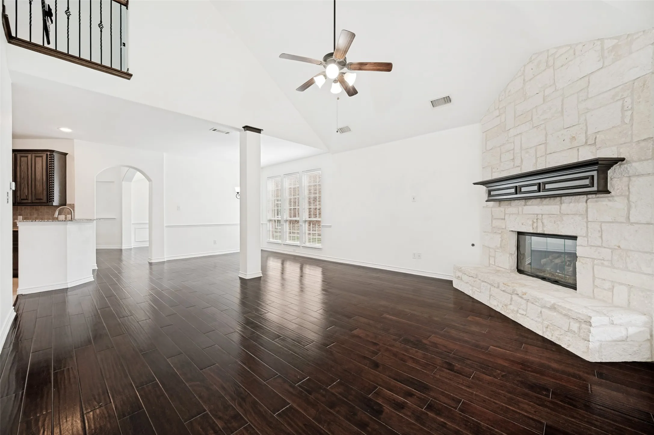Spacious living area featuring high vaulted ceilings with a ceiling fan, dark wood-finish flooring, and a stone-clad fireplace with a dark wood mantel
