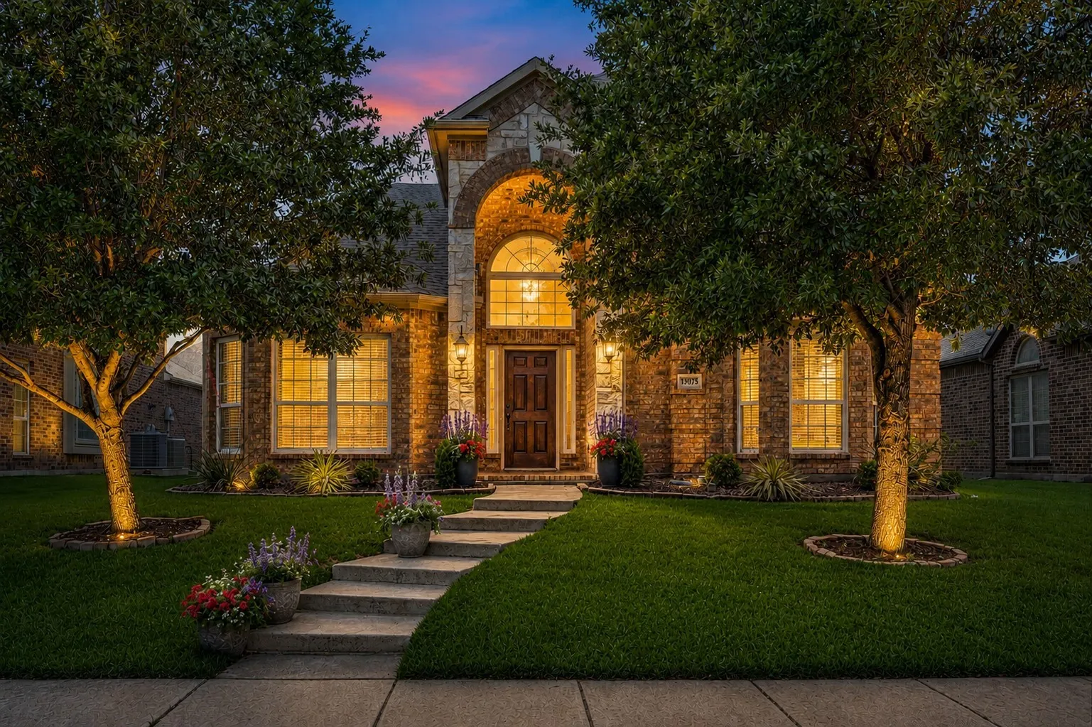 Brick and stone facade featuring a grand arched entryway, multi-paned windows, a well-maintained lawn, mature trees with uplighting, and a concrete pathway leading to the front door.
AI enhanced