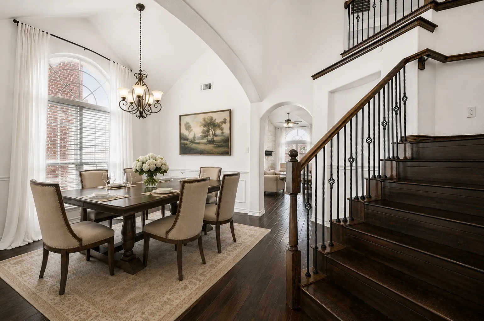 Formal dining area featuring a vaulted ceiling with an ornate chandelier, a large arched window with blinds, and rich wood-finish flooring.
Virtually Staged