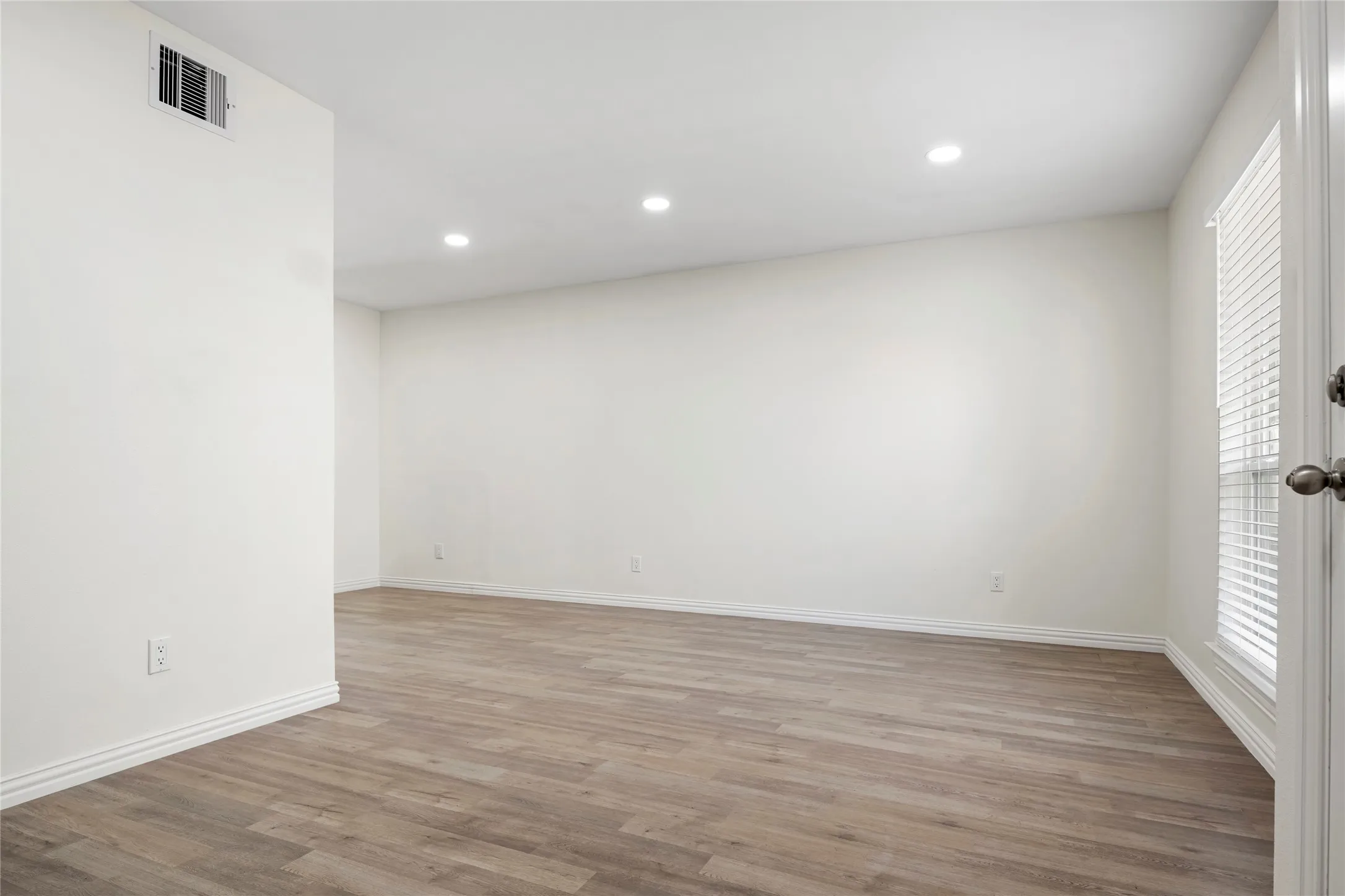 Spacious room featuring wood-finish flooring, white baseboards, recessed ceiling lighting, and a window with blinds