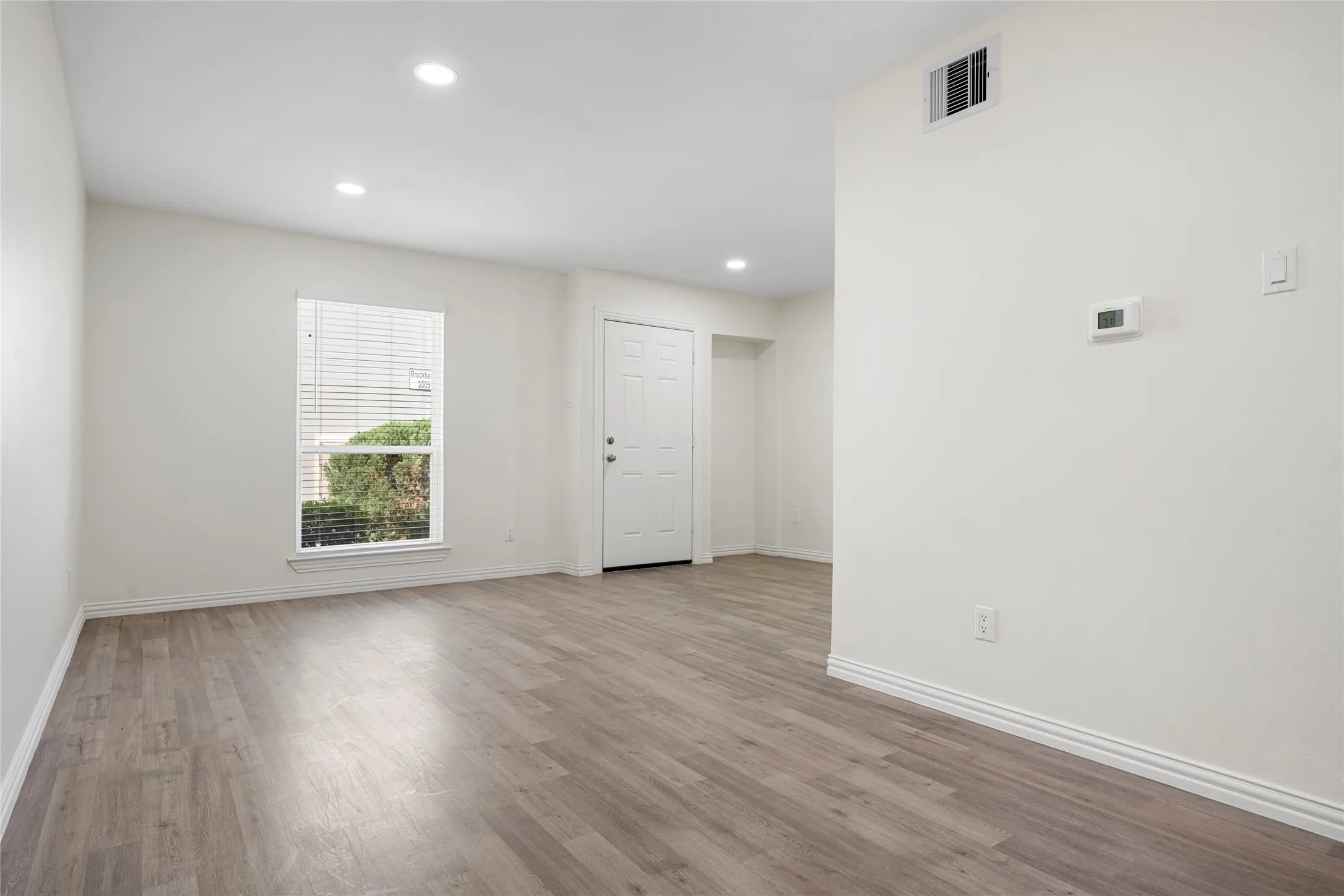 Spacious interior featuring wood-finish flooring, recessed lighting, a window with blinds, a paneled entry door, and a digital thermostat