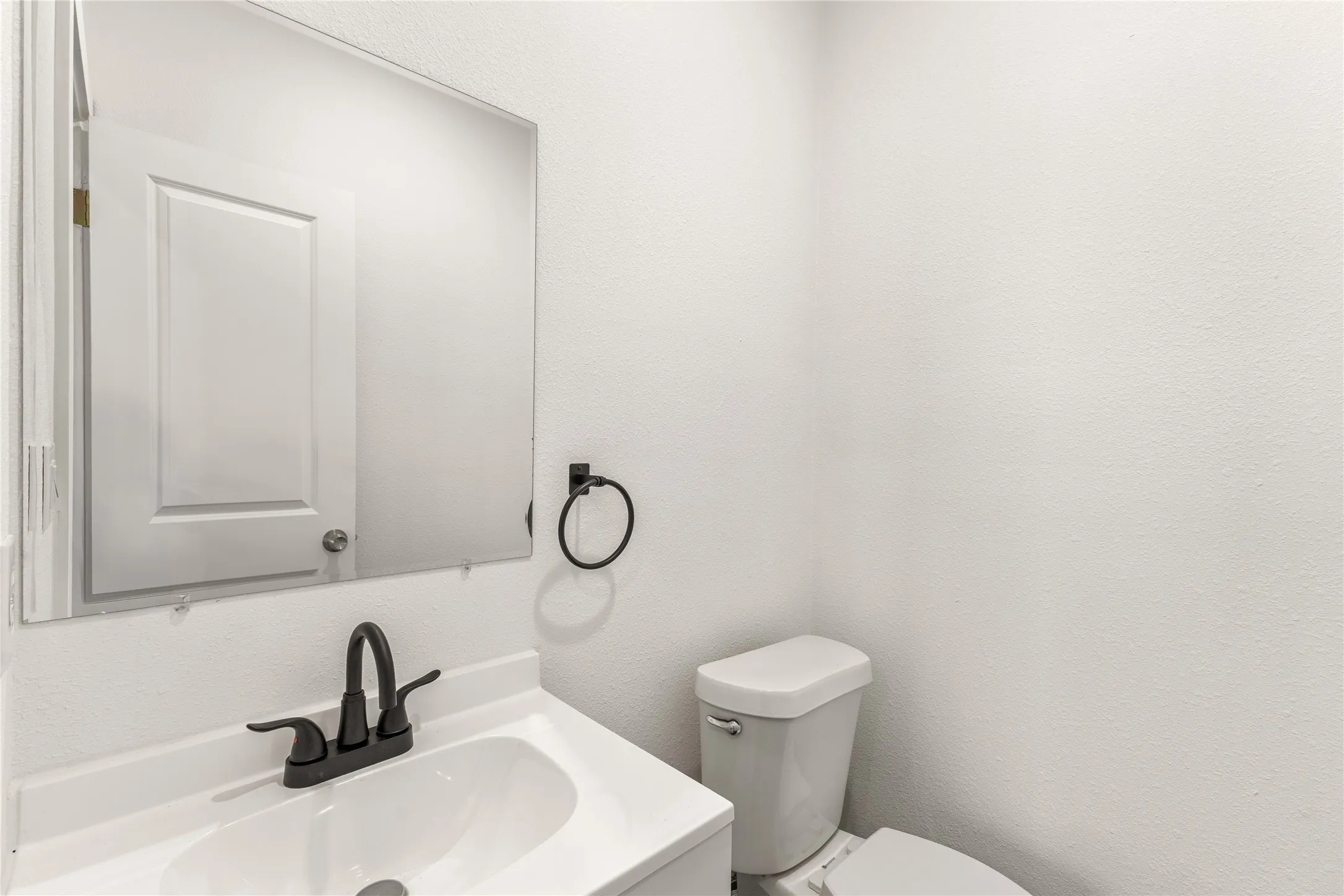 Bathroom featuring a white vanity with integrated sink, black faucet, wall-mounted mirror, and white commode