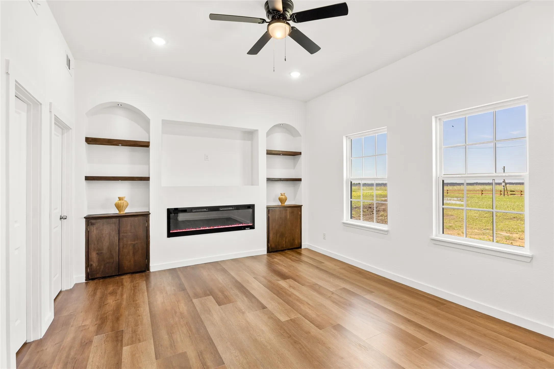 Spacious room featuring wood-finish flooring, an electric fireplace with an integrated media niche, built-in shelving with arched alcoves, and two double-hung windows
