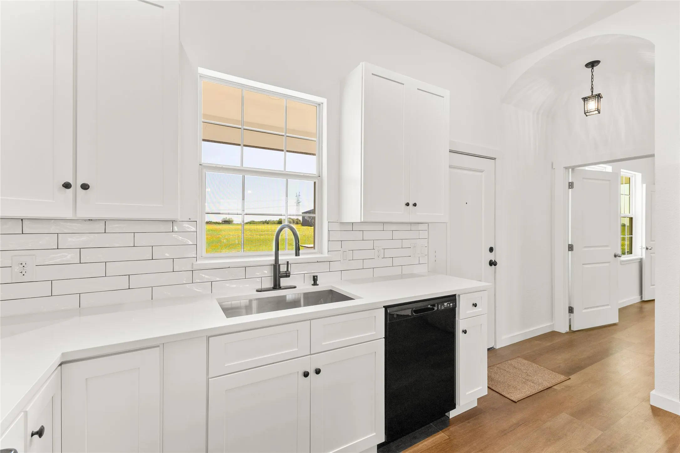 Kitchen featuring white shaker-style cabinetry, white subway tile backsplash, stainless steel double-basin sink with a matte black gooseneck faucet, white solid surface countertops, and wood-finish flooring