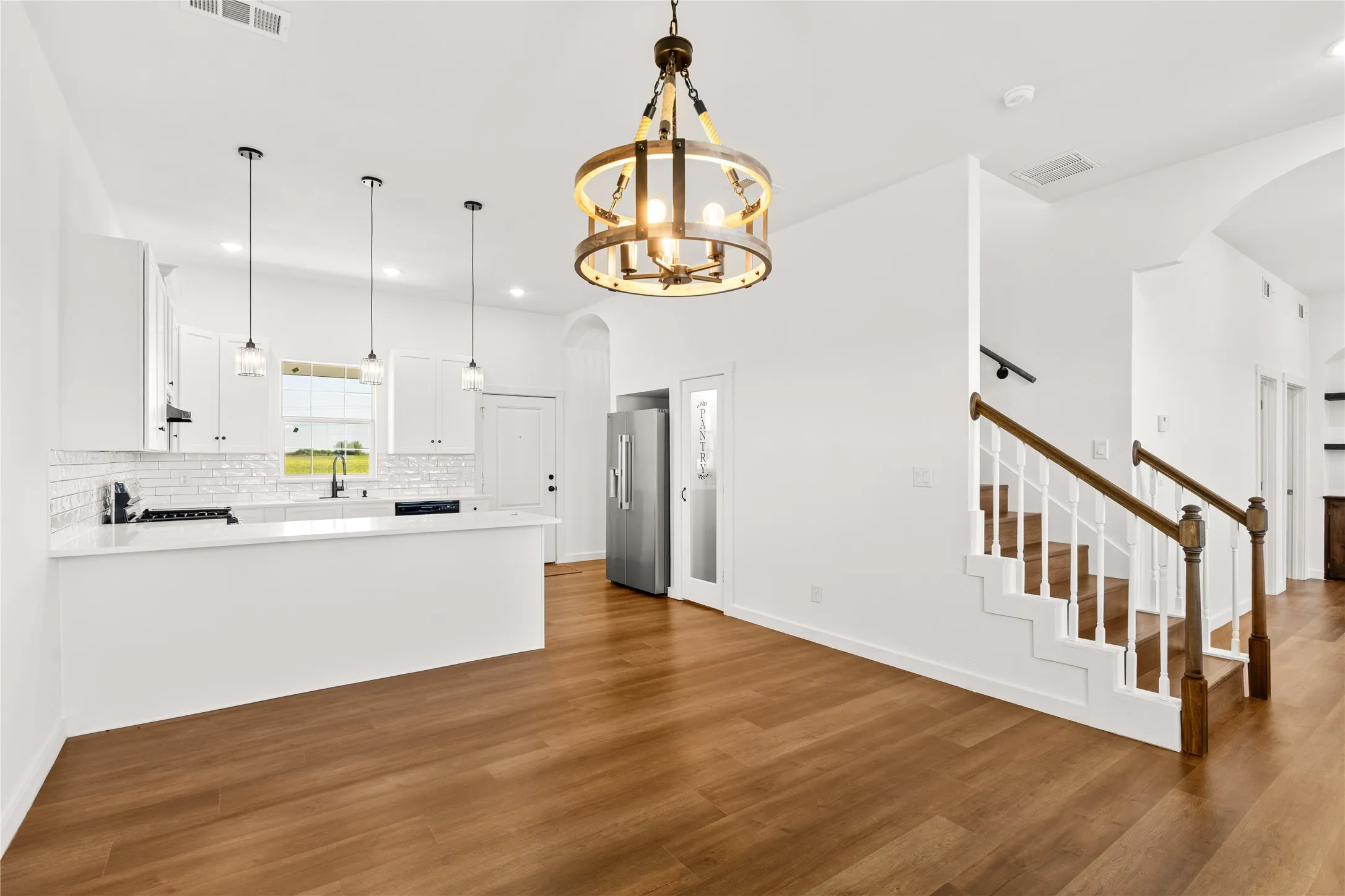 Open-concept living space featuring wood-finish flooring, a contemporary kitchen with white cabinetry and subway tile backsplash, and a staircase with a white baluster and wood handrail