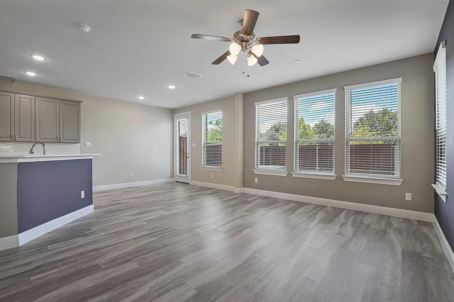 Expansive open concept living area featuring wood-finish flooring, a contemporary ceiling fan, and multiple windows with horizontal blinds